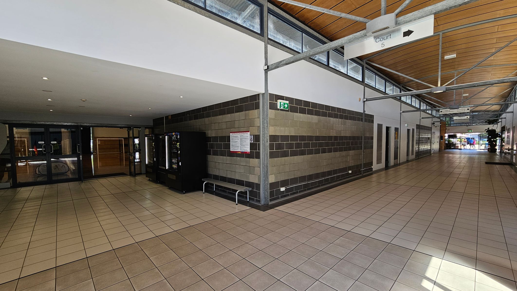 A corridor with a tiled floor and a wooden panelled ceiling. On the left, there are glass doors leading to another area, with vending machines nearby. The right side features a wall with a striped pattern of grey and beige bricks, a bench, and an emergency exit sign. Further down the corridor, signs hang from the ceiling, and there is a glimpse of people near the entrance at the far end. A corridor with a tiled floor and a wooden panelled ceiling. On the left, there are glass doors leading to another area, with vending machines nearby. The right side features a wall with a striped pattern of grey and beige bricks, a bench, and an emergency exit sign. Further down the corridor, signs hang from the ceiling, and there is a glimpse of people near the entrance at the far end.