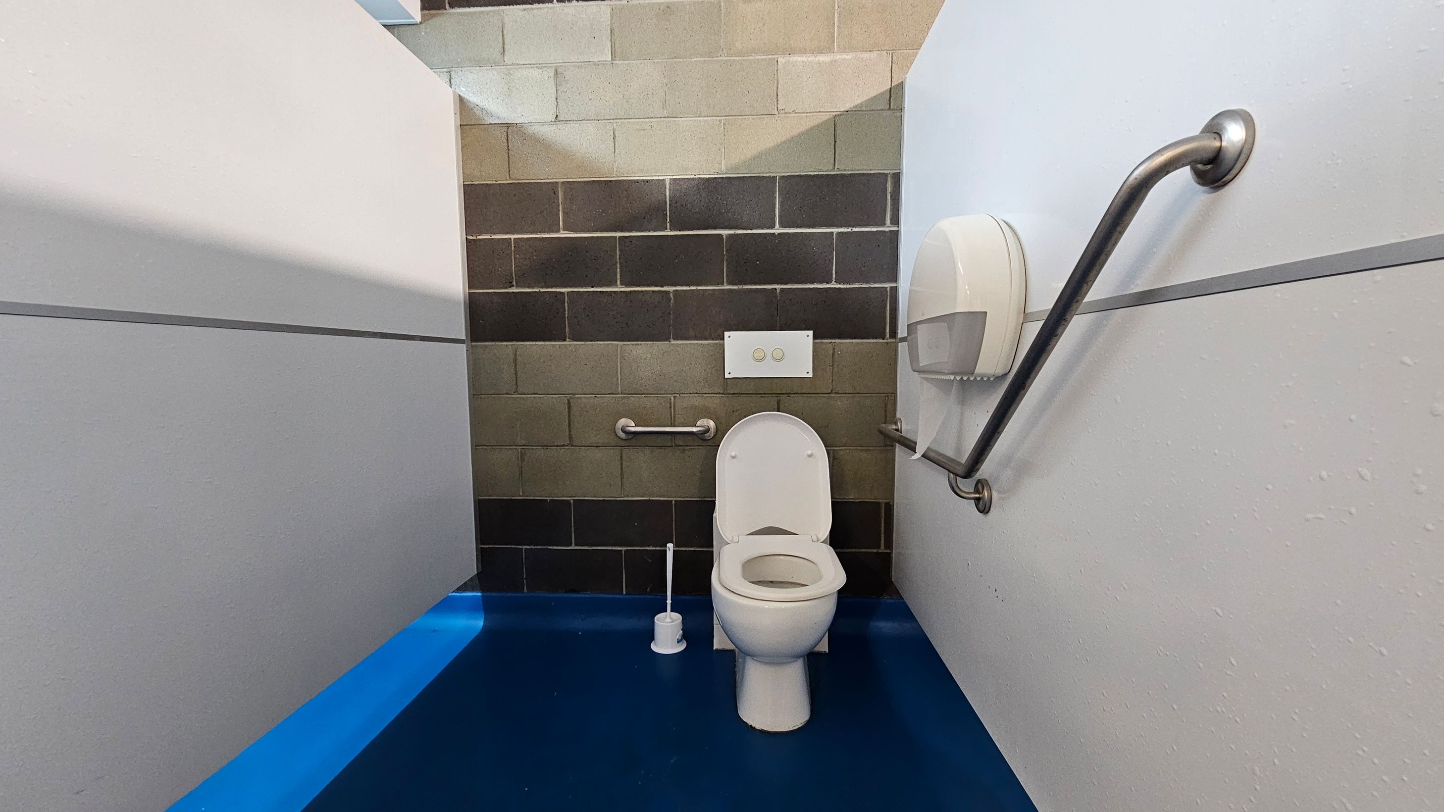 A public toilet with a white toilet bowl and seat in the centre. The back wall is made of multicoloured bricks. There are two metal grab bars on either side of the toilet, with a toilet paper dispenser on the right. The floor is a bright blue, and the walls are white with a horizontal dividing line. A toilet brush is placed on the floor to the left of the toilet. A public toilet with a white toilet bowl and seat in the centre. The back wall is made of multicoloured bricks. There are two metal grab bars on either side of the toilet, with a toilet paper dispenser on the right. The floor is a bright blue, and the walls are white with a horizontal dividing line. A toilet brush is placed on the floor to the left of the toilet.