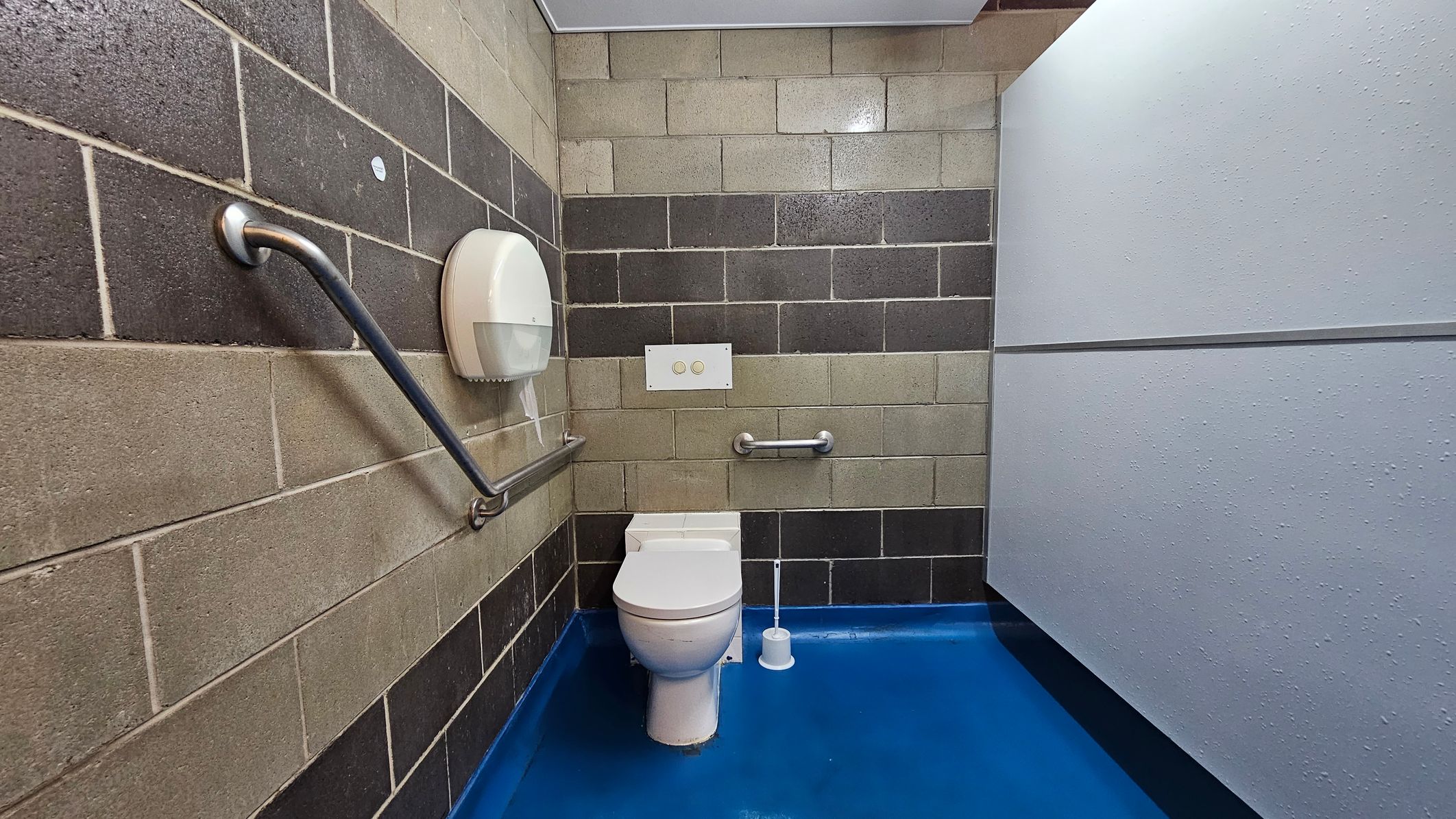 A public toilet with a blue floor and a white ceiling. The walls are made of grey and beige concrete blocks. On the left, there's a metal grab bar and a toilet paper dispenser. In the centre, a white toilet with a dual-flush button panel above it and another grab bar to the right. A toilet brush is placed on the floor next to the toilet. On the right, a light grey partition wall. A public toilet with a blue floor and a white ceiling. The walls are made of grey and beige concrete blocks. On the left, there's a metal grab bar and a toilet paper dispenser. In the centre, a white toilet with a dual-flush button panel above it and another grab bar to the right. A toilet brush is placed on the floor next to the toilet. On the right, a light grey partition wall.