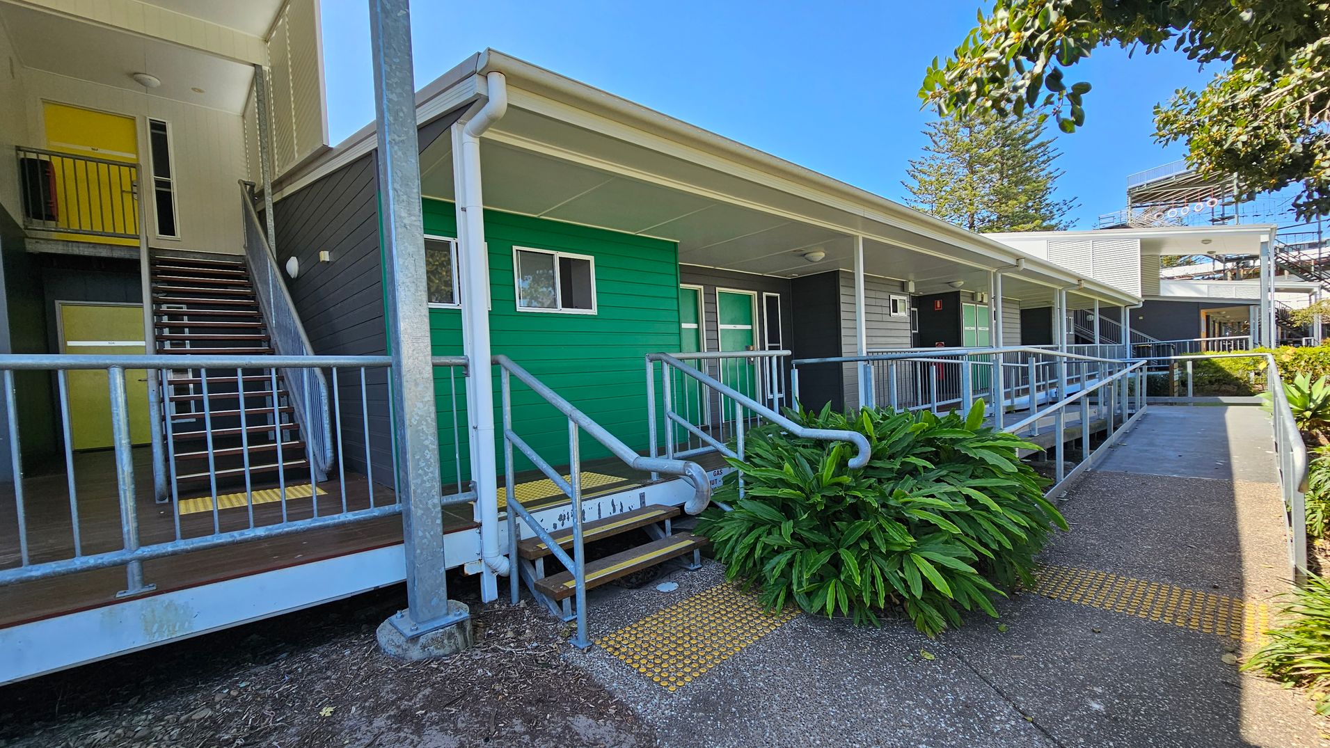 A building with a green exterior wall and a covered walkway. On the left, there is a staircase with a metal railing leading to an upper level with a yellow door. The foreground features a ramp with handrails and tactile paving. A lush green plant is near the ramp. The building has a flat roof and a grey section further along. Trees and a structure with a metal framework are visible in the background. The ground is paved with a textured surface. A building with a green exterior wall and a covered walkway. On the left, there is a staircase with a metal railing leading to an upper level with a yellow door. The foreground features a ramp with handrails and tactile paving. A lush green plant is near the ramp. The building has a flat roof and a grey section further along. Trees and a structure with a metal framework are visible in the background. The ground is paved with a textured surface.