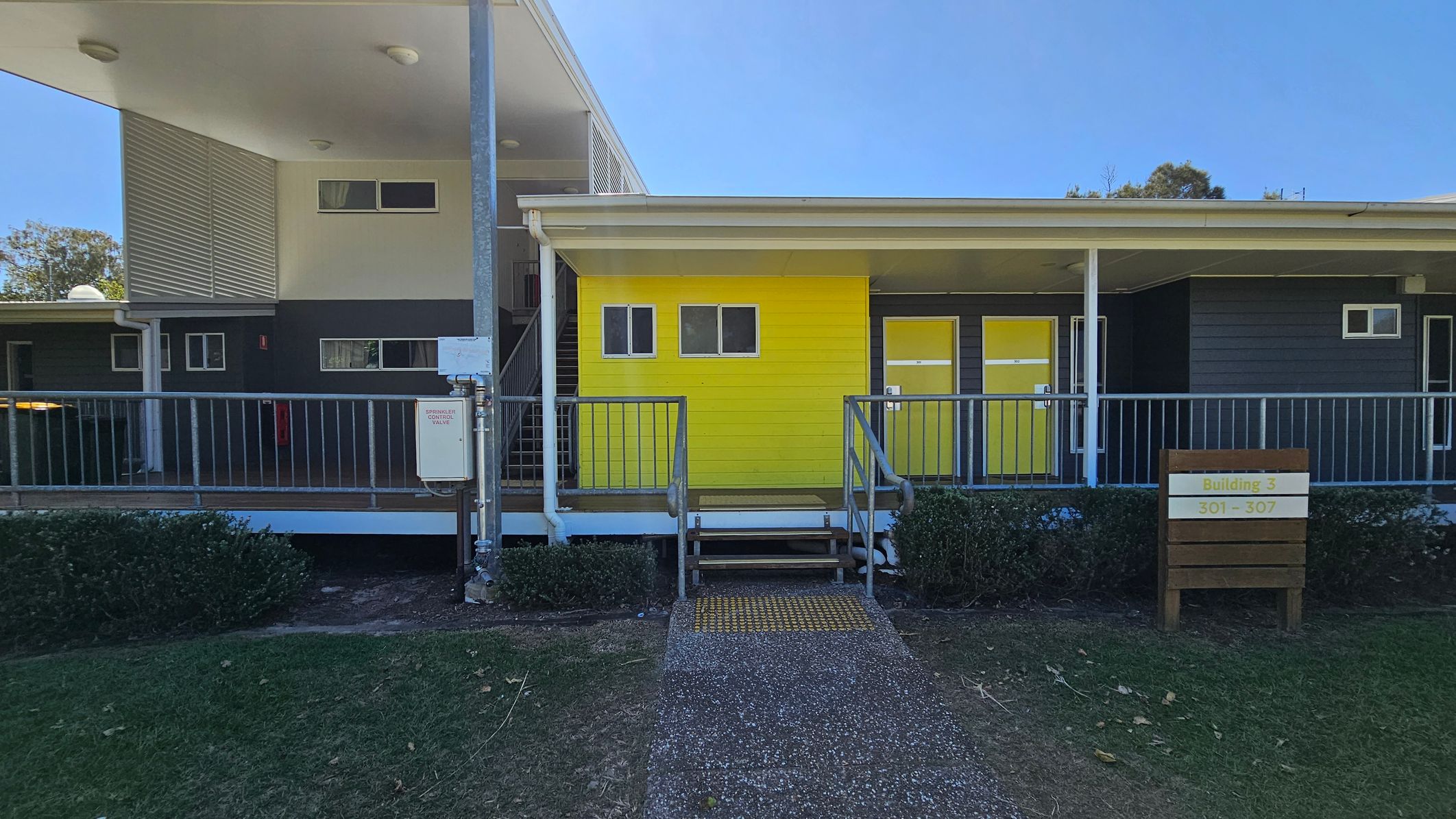 A building with a bright yellow section in the centre, featuring two small windows and a door. To the right, there is a sign reading "Building 3, 301 - 307" and another yellow door. The left side has a grey section with a staircase and additional windows. A metal railing runs along the front, with a small set of stairs leading up to the entrance. The foreground has a path and green grass, with bushes lining the building. The ceiling is white with recessed lights. A building with a bright yellow section in the centre, featuring two small windows and a door. To the right, there is a sign reading "Building 3, 301 - 307" and another yellow door. The left side has a grey section with a staircase and additional windows. A metal railing runs along the front, with a small set of stairs leading up to the entrance. The foreground has a path and green grass, with bushes lining the building. The ceiling is white with recessed lights.
