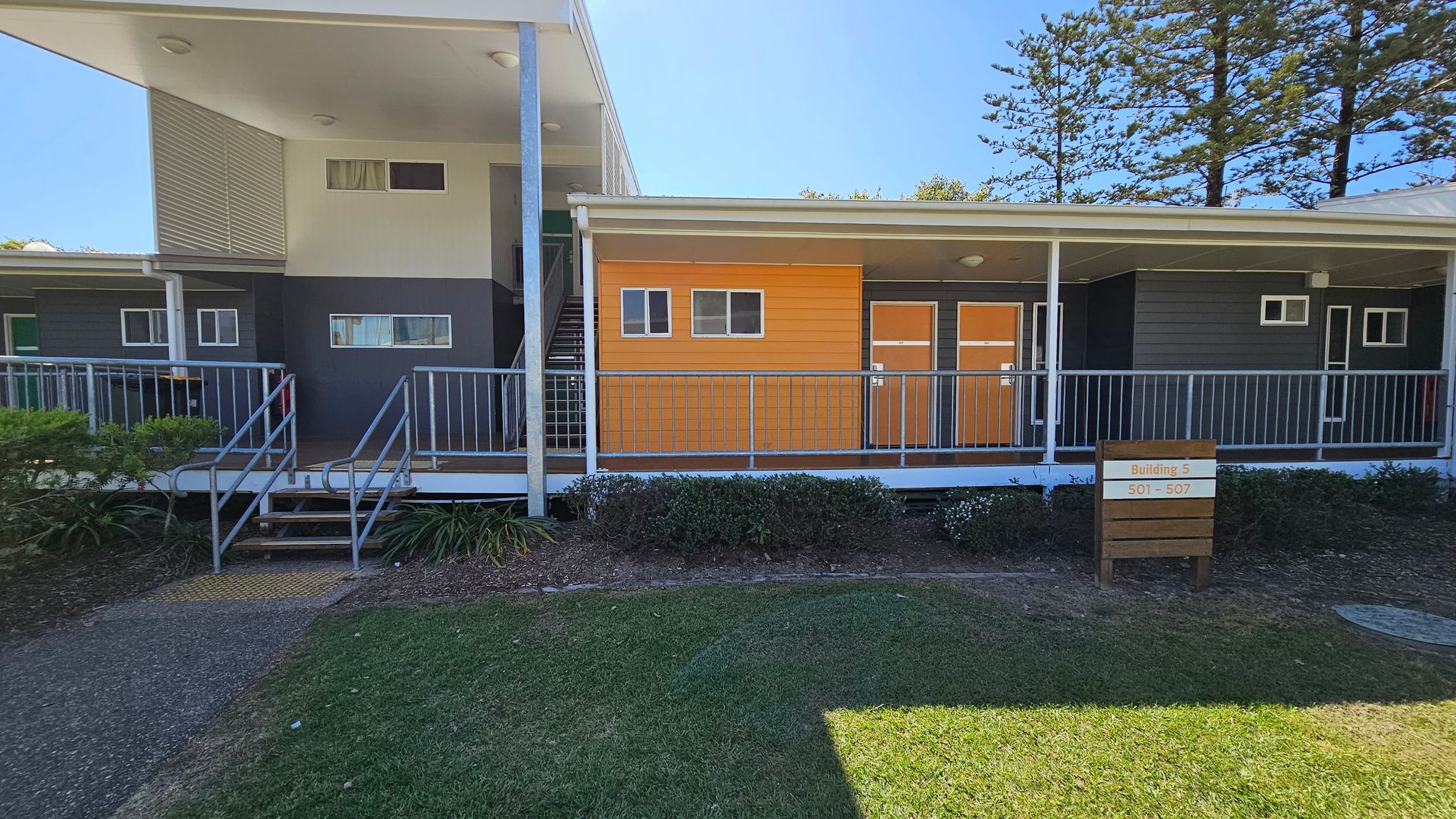 A modern building with a flat roof and a mix of grey and orange panelling. On the left, there is a staircase leading to an upper level. The building has several small rectangular windows. In front, a metal railing runs along a walkway. A wooden sign in the foreground reads "Building 5, 501 - 507". The ground is covered with grass and shrubs, and tall trees are visible in the background. A modern building with a flat roof and a mix of grey and orange panelling. On the left, there is a staircase leading to an upper level. The building has several small rectangular windows. In front, a metal railing runs along a walkway. A wooden sign in the foreground reads "Building 5, 501 - 507". The ground is covered with grass and shrubs, and tall trees are visible in the background.