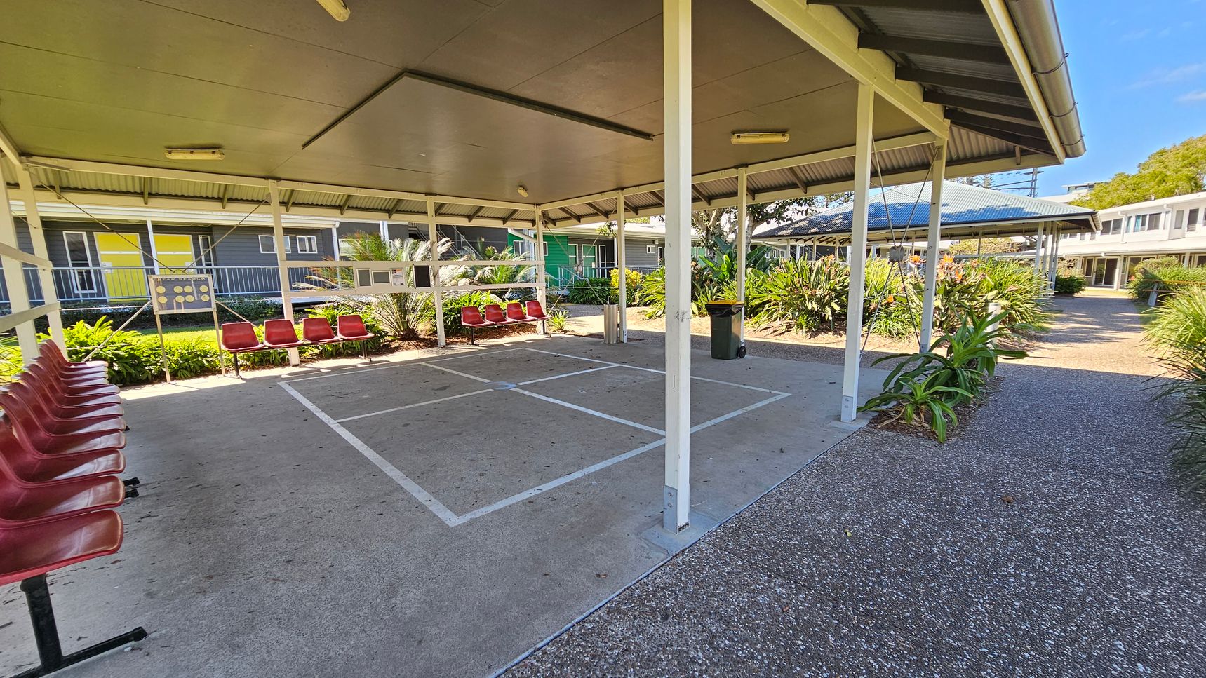 A covered outdoor area with a concrete floor marked with white lines. Red plastic chairs are lined up on the left side. The structure has a metal roof supported by white poles. Surrounding the area are green plants and shrubs. In the background, there are buildings with yellow and green walls, and a pathway with a speckled surface leads through the space. A rubbish bin is visible near the centre. A covered outdoor area with a concrete floor marked with white lines. Red plastic chairs are lined up on the left side. The structure has a metal roof supported by white poles. Surrounding the area are green plants and shrubs. In the background, there are buildings with yellow and green walls, and a pathway with a speckled surface leads through the space. A rubbish bin is visible near the centre.