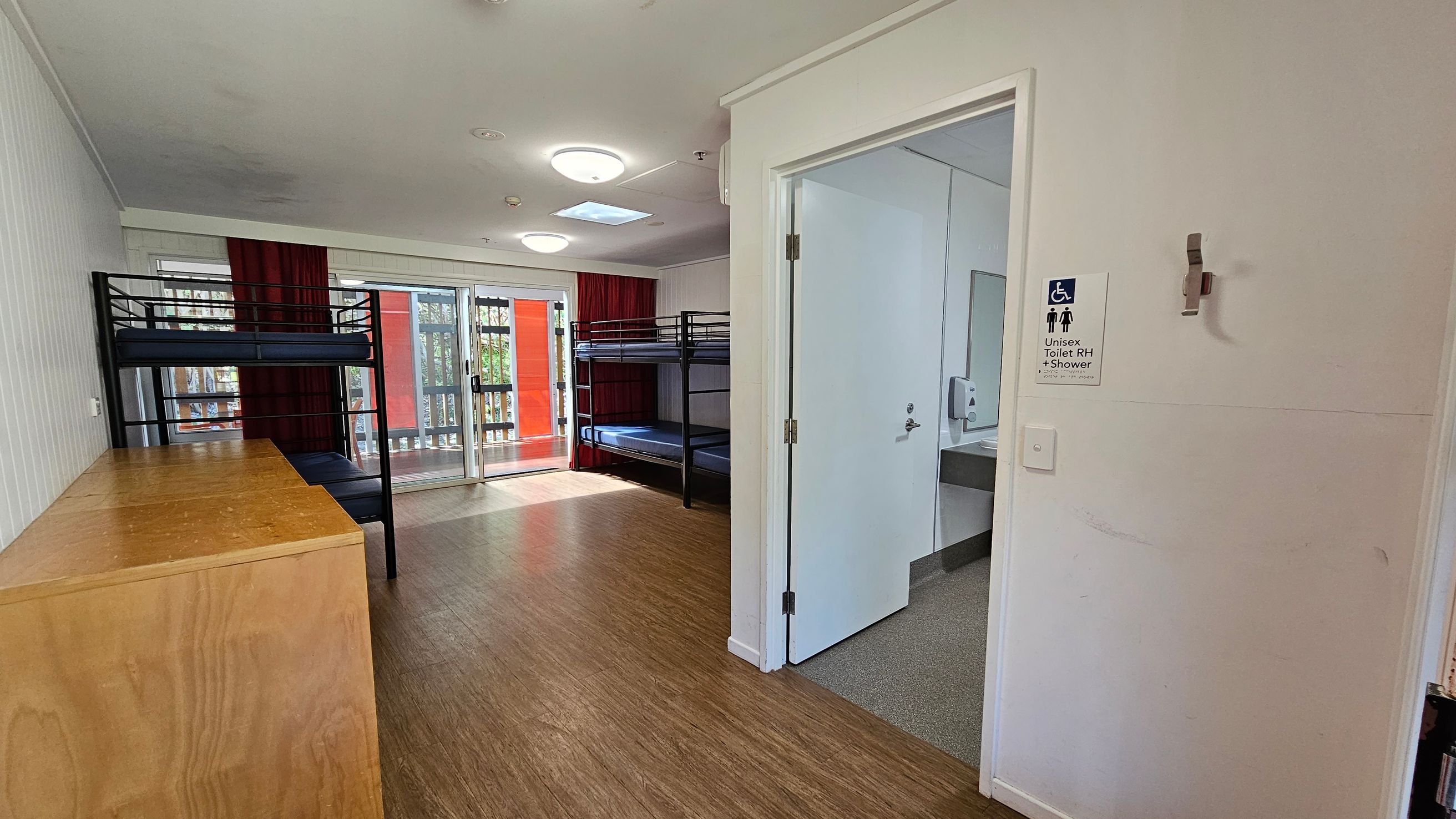 A room with wooden flooring and a white ceiling. On the left, there are two sets of metal bunk beds and a wooden cabinet. In the centre, a sliding glass door with red curtains leads to a balcony. On the right, an open door reveals a bathroom with a sign indicating it is a unisex toilet with a shower. The bathroom has a sink and a soap dispenser visible. A room with wooden flooring and a white ceiling. On the left, there are two sets of metal bunk beds and a wooden cabinet. In the centre, a sliding glass door with red curtains leads to a balcony. On the right, an open door reveals a bathroom with a sign indicating it is a unisex toilet with a shower. The bathroom has a sink and a soap dispenser visible.