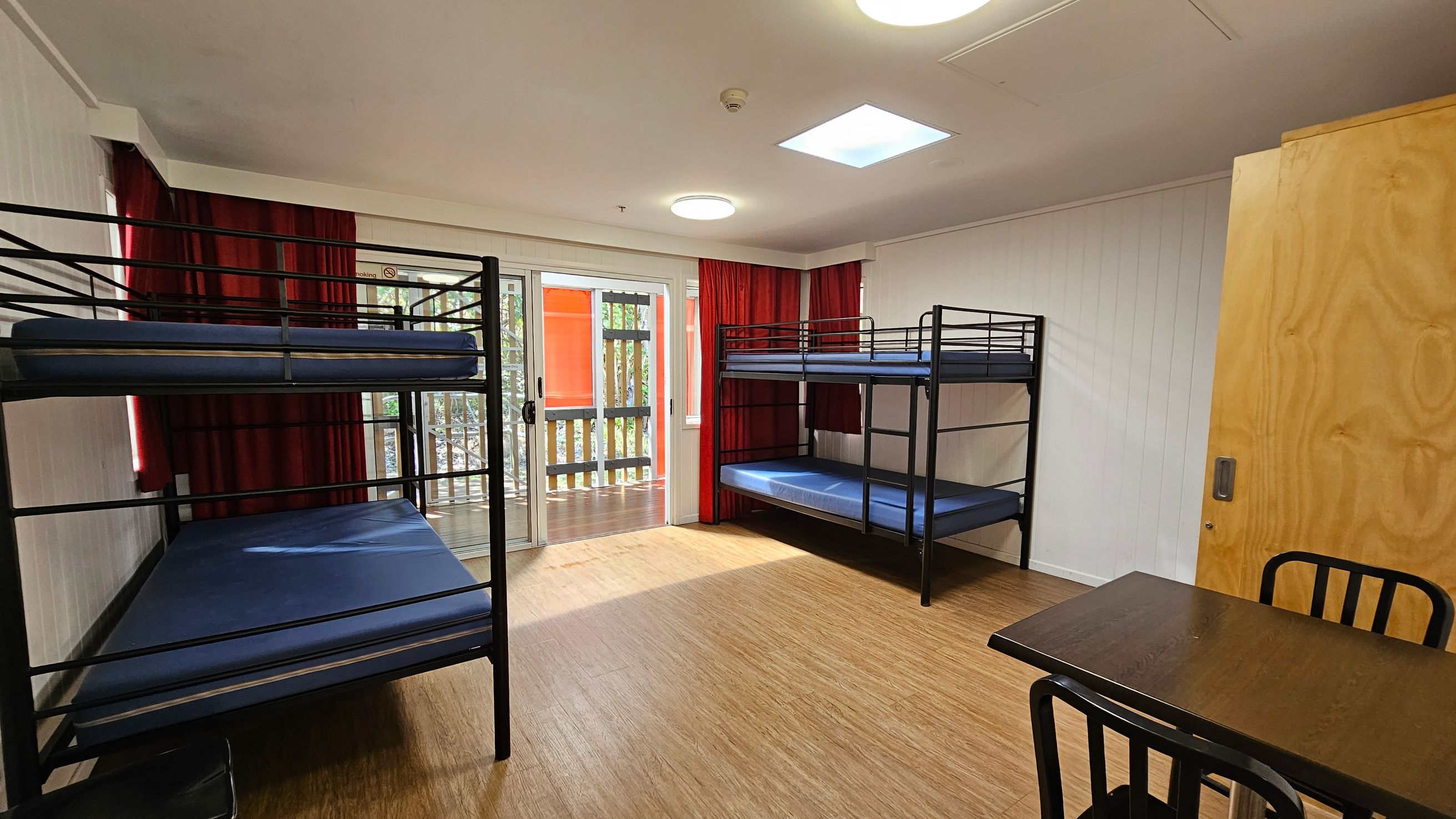 A room with two sets of black metal bunk beds, each with blue mattresses, positioned on the left and right sides. In the centre, there are large sliding glass doors with red curtains, leading to a balcony with a wooden railing. On the right, a wooden door and a small black table with chairs are visible. The floor is light wood, and the ceiling has recessed lighting and a skylight. A room with two sets of black metal bunk beds, each with blue mattresses, positioned on the left and right sides. In the centre, there are large sliding glass doors with red curtains, leading to a balcony with a wooden railing. On the right, a wooden door and a small black table with chairs are visible. The floor is light wood, and the ceiling has recessed lighting and a skylight.