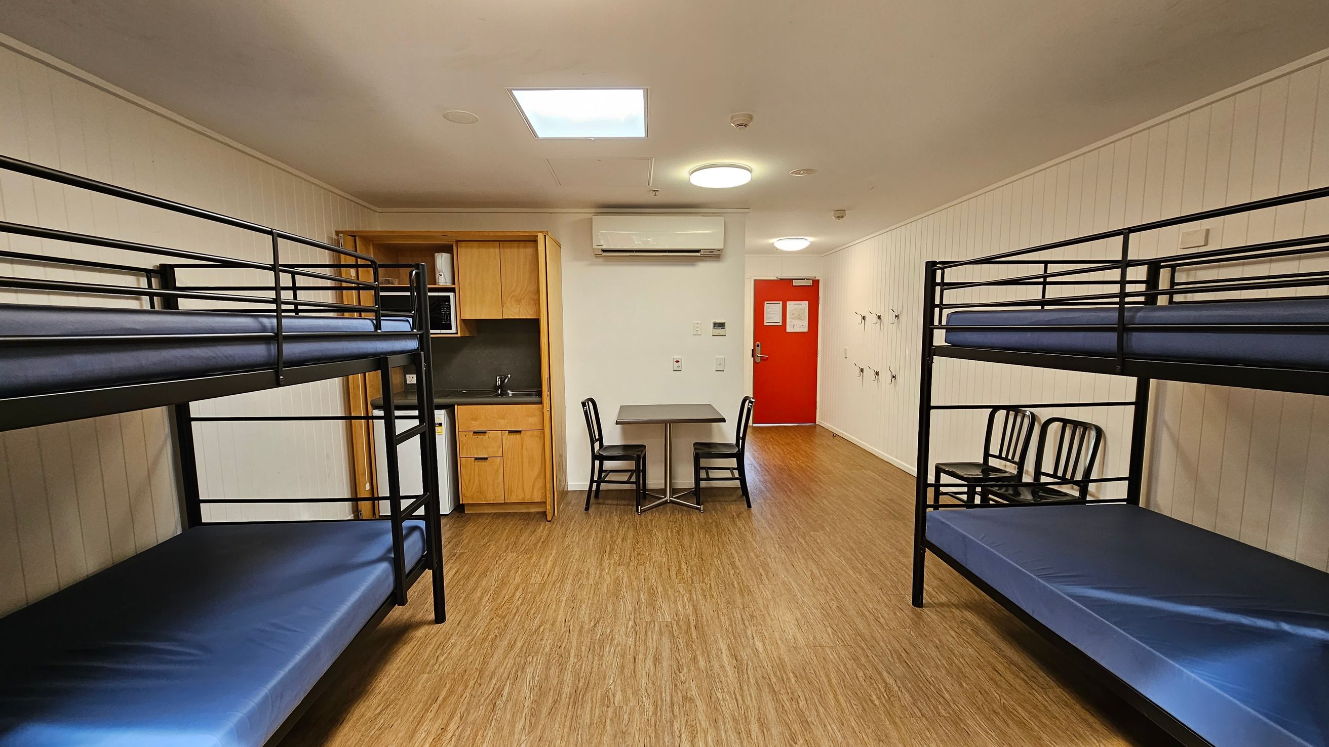A room with two sets of metal bunk beds on the left and right, each with blue mattresses. In the centre, there's a small table with two black chairs. The back wall features a red door and a wall-mounted air conditioning unit above the table. To the left, there's a kitchenette with wooden cabinets, a microwave, and a small sink. The floor is wooden, and the walls are white with vertical panelling. Ceiling lights are visible above. A room with two sets of metal bunk beds on the left and right, each with blue mattresses. In the centre, there's a small table with two black chairs. The back wall features a red door and a wall-mounted air conditioning unit above the table. To the left, there's a kitchenette with wooden cabinets, a microwave, and a small sink. The floor is wooden, and the walls are white with vertical panelling. Ceiling lights are visible above.