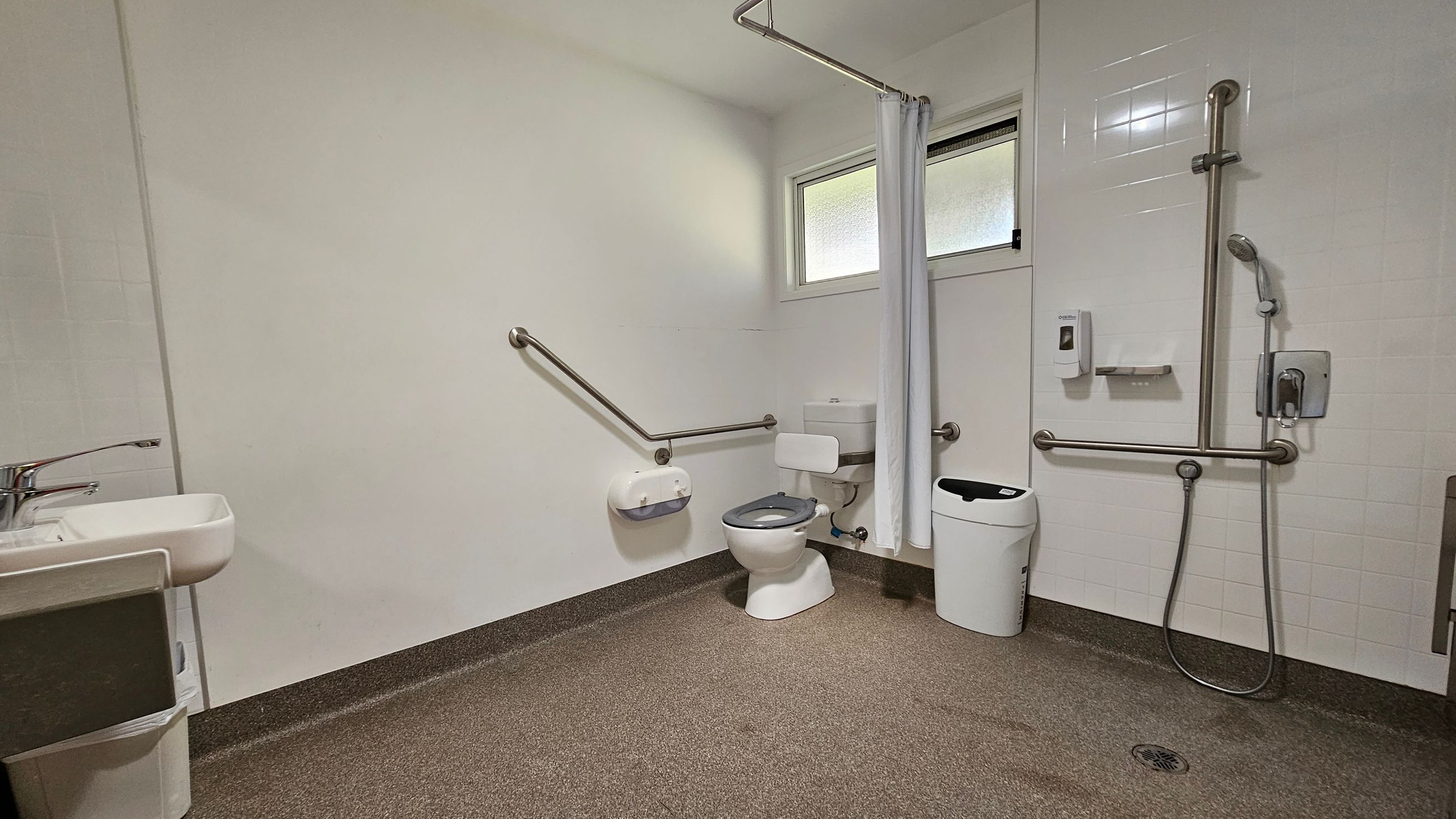 A spacious, accessible bathroom with a non-slip brown floor and light-coloured walls. On the left, there's a sink with a single lever tap. In the centre, a toilet with grab bars on both sides and a toilet paper holder. To the right, a shower area with a handheld showerhead and additional grab bars. A white curtain separates the shower from the rest of the room. A small window is above the toilet, and a waste bin is nearby. A spacious, accessible bathroom with a non-slip brown floor and light-coloured walls. On the left, there's a sink with a single lever tap. In the centre, a toilet with grab bars on both sides and a toilet paper holder. To the right, a shower area with a handheld showerhead and additional grab bars. A white curtain separates the shower from the rest of the room. A small window is above the toilet, and a waste bin is nearby.