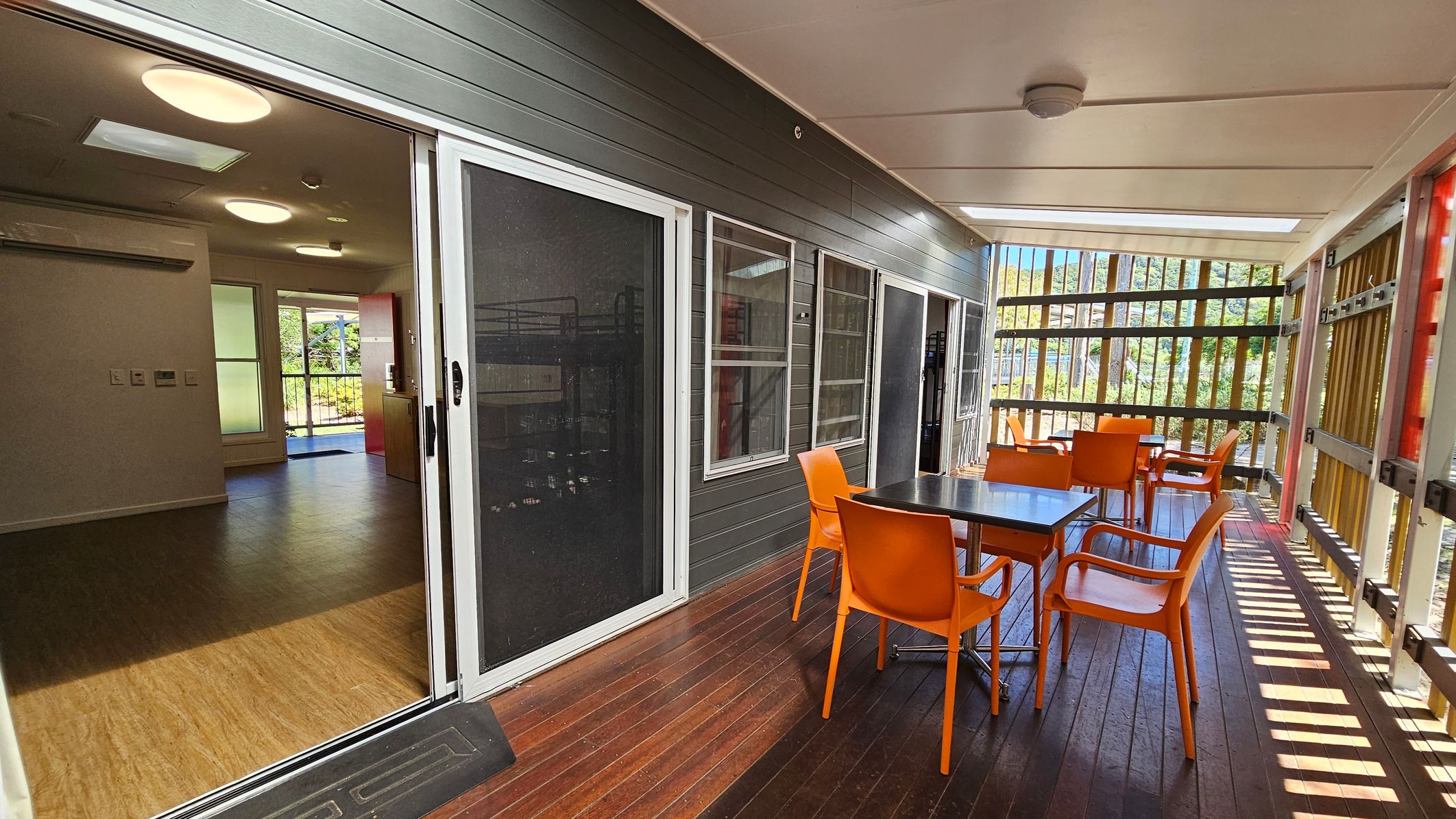 A covered outdoor area with wooden flooring and a ceiling with recessed lighting. On the right, there is a table with four bright orange chairs. The left side features sliding glass doors leading to an interior space with wooden flooring and ceiling lights. The background shows vertical wooden slats allowing natural light to filter through, with greenery visible beyond. A covered outdoor area with wooden flooring and a ceiling with recessed lighting. On the right, there is a table with four bright orange chairs. The left side features sliding glass doors leading to an interior space with wooden flooring and ceiling lights. The background shows vertical wooden slats allowing natural light to filter through, with greenery visible beyond.