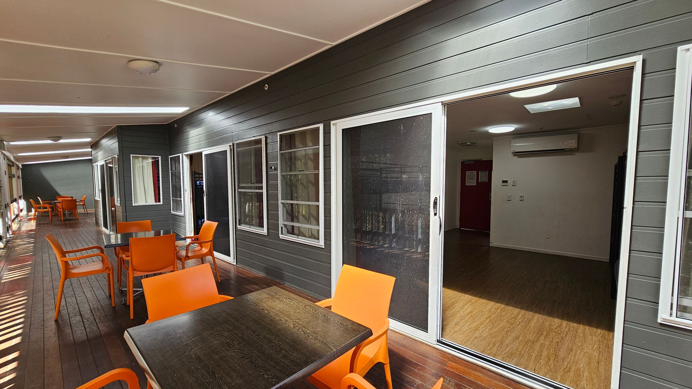A covered outdoor area with a wooden floor and a white ceiling. On the left, there are several tables with bright orange chairs. The right side features a dark grey wall with multiple windows and a sliding glass door leading to an interior room. The room inside has a wooden floor, white walls, and a visible air conditioning unit. The space is well-lit with ceiling lights. A covered outdoor area with a wooden floor and a white ceiling. On the left, there are several tables with bright orange chairs. The right side features a dark grey wall with multiple windows and a sliding glass door leading to an interior room. The room inside has a wooden floor, white walls, and a visible air conditioning unit. The space is well-lit with ceiling lights.