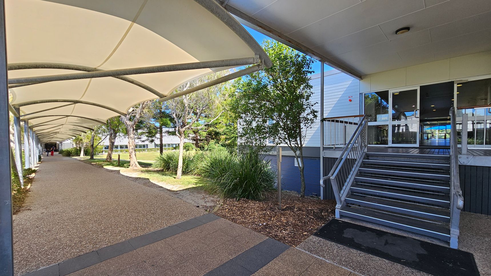A covered walkway on the left with a curved, white canopy supported by metal beams. The path is paved with a textured surface. On the right, there is a building with a set of metal stairs leading to glass doors. Trees and shrubs are planted along the side of the building, and a grassy area is visible in the background. The ceiling of the walkway is made of fabric, and the building has a flat, white ceiling. A covered walkway on the left with a curved, white canopy supported by metal beams. The path is paved with a textured surface. On the right, there is a building with a set of metal stairs leading to glass doors. Trees and shrubs are planted along the side of the building, and a grassy area is visible in the background. The ceiling of the walkway is made of fabric, and the building has a flat, white ceiling.