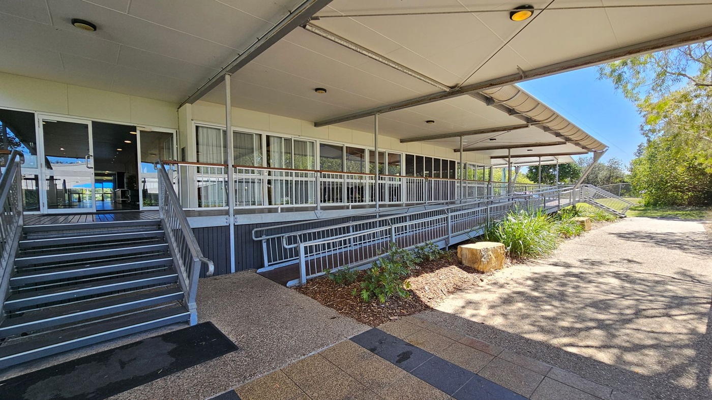 A building entrance with a covered walkway. On the left, there are metal stairs leading up to glass doors. The middle section features a long ramp with railings, bordered by a small garden with plants and a large stone. On the right, a paved path extends alongside the building, surrounded by trees and greenery. The ceiling is flat with recessed lights, and the floor is a mix of textured and smooth surfaces. A building entrance with a covered walkway. On the left, there are metal stairs leading up to glass doors. The middle section features a long ramp with railings, bordered by a small garden with plants and a large stone. On the right, a paved path extends alongside the building, surrounded by trees and greenery. The ceiling is flat with recessed lights, and the floor is a mix of textured and smooth surfaces.