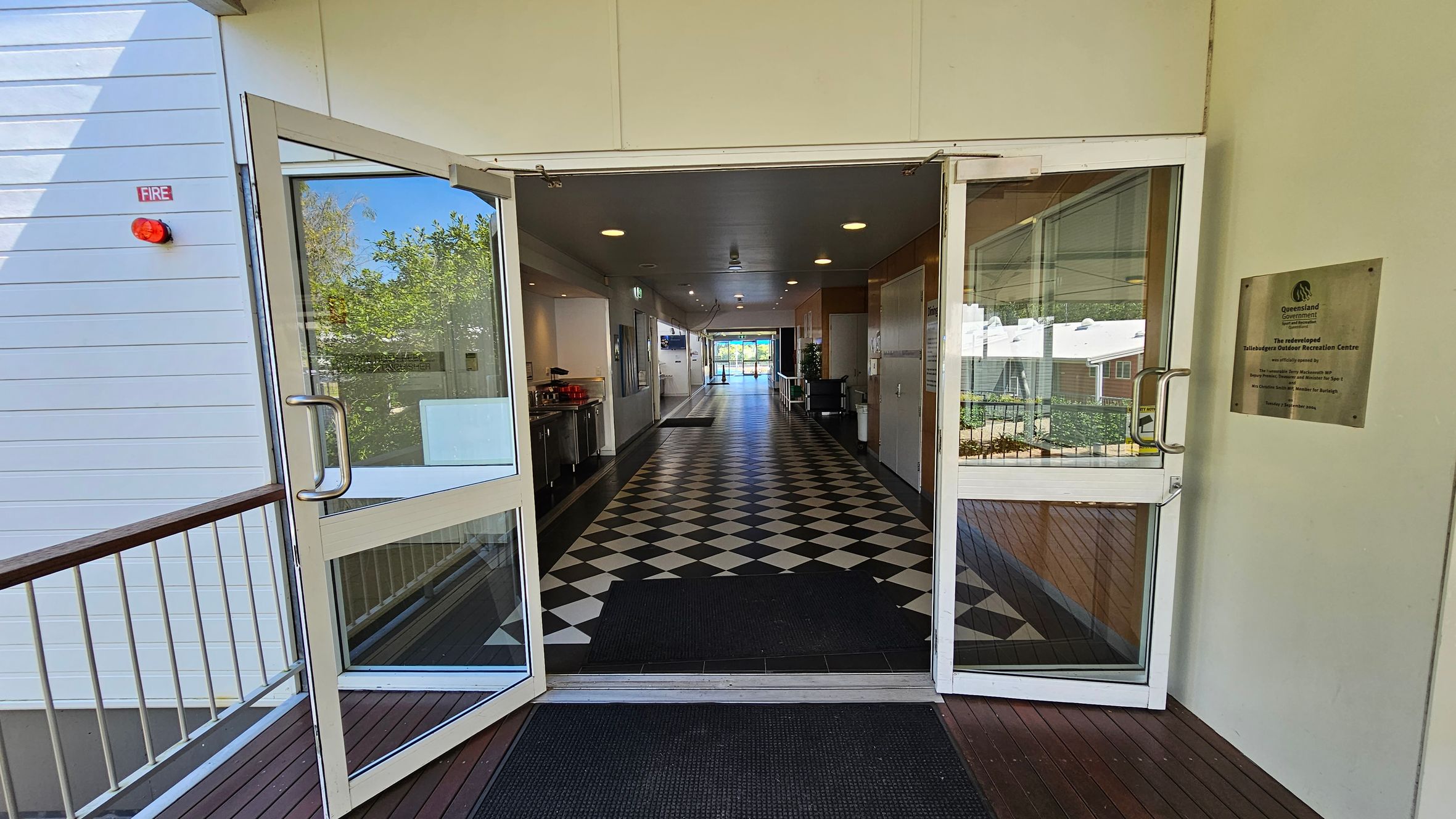 Glass double doors open into a long corridor with a black and white chequered floor. On the left, there's a small kitchen area with a counter and appliances. On the right, a plaque is mounted on the wall. The corridor is well-lit with ceiling lights and leads to a bright area with large windows at the far end. Outside the doors, a wooden deck with metal railings is visible, along with some greenery. Glass double doors open into a long corridor with a black and white chequered floor. On the left, there's a small kitchen area with a counter and appliances. On the right, a plaque is mounted on the wall. The corridor is well-lit with ceiling lights and leads to a bright area with large windows at the far end. Outside the doors, a wooden deck with metal railings is visible, along with some greenery.