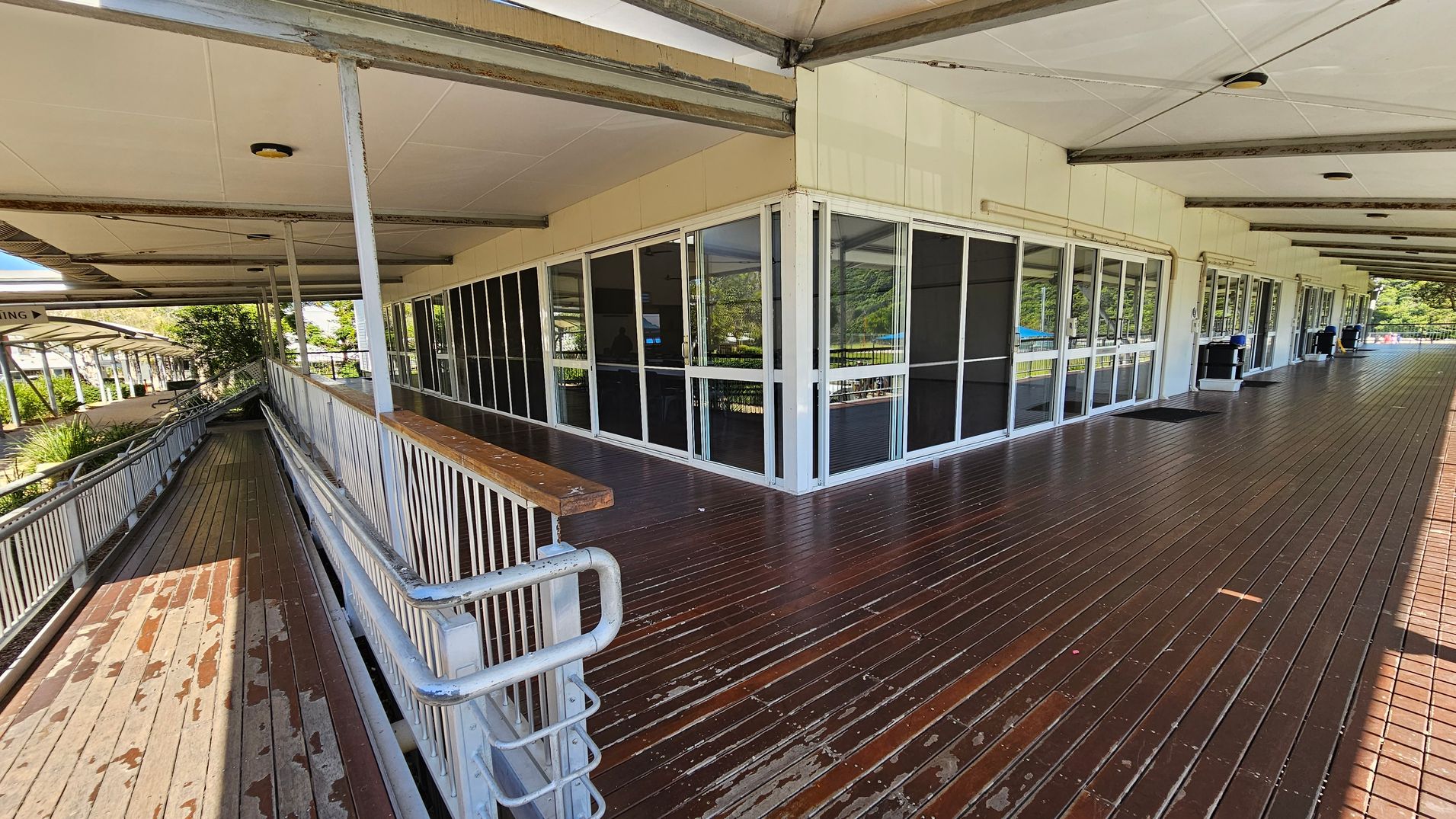 A wooden deck with a ramp on the left side, featuring metal railings. The building has large glass windows and doors with dark panels. The ceiling is supported by metal beams, and there are several ceiling lights. In the background, there are trees and a covered walkway. The floor is made of dark wooden planks, and there are a few scattered items on it. A wooden deck with a ramp on the left side, featuring metal railings. The building has large glass windows and doors with dark panels. The ceiling is supported by metal beams, and there are several ceiling lights. In the background, there are trees and a covered walkway. The floor is made of dark wooden planks, and there are a few scattered items on it.