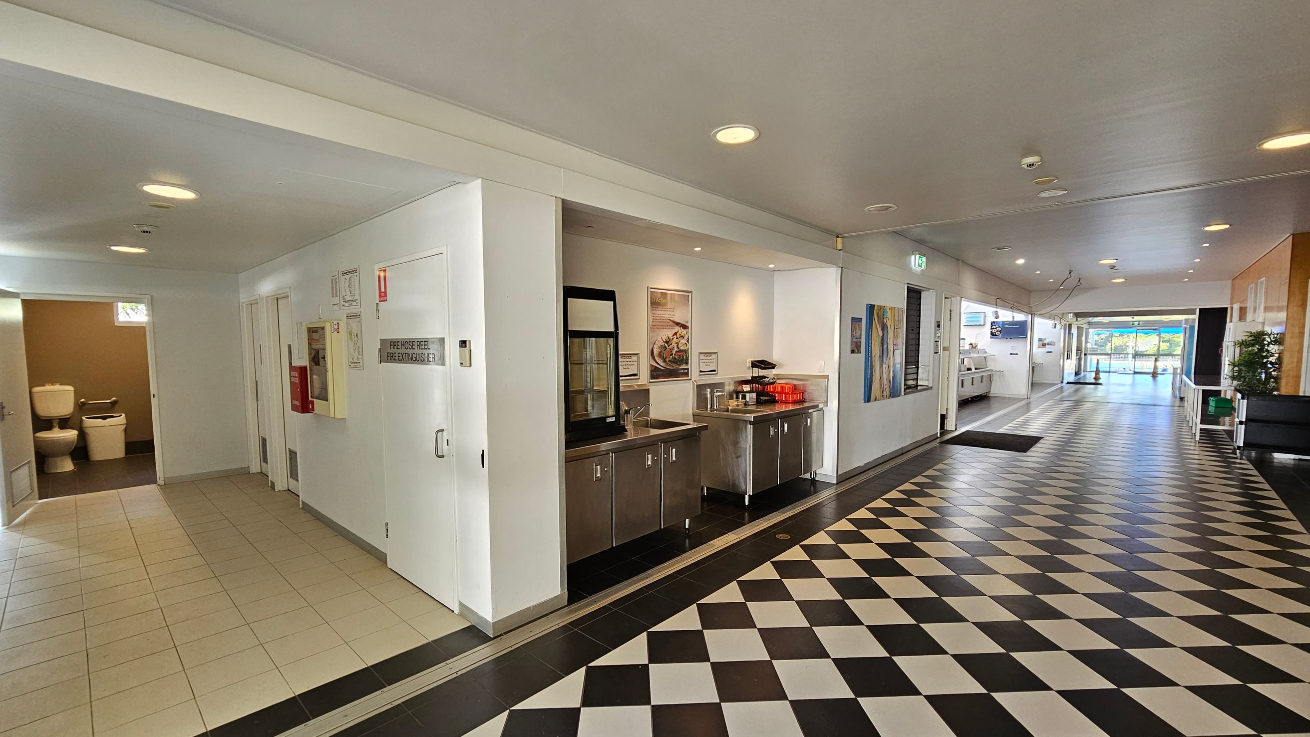A corridor with a black and white chequered floor. On the left, there is a tiled area leading to a bathroom with visible toilets. Adjacent to this is a wall with a fire hose reel and extinguisher cabinet. In the middle, a stainless steel counter with a sink and red trays is set against the wall. The right side of the corridor has a series of artworks and leads to a glass door with a view outside. The ceiling is white with recessed lighting. A corridor with a black and white chequered floor. On the left, there is a tiled area leading to a bathroom with visible toilets. Adjacent to this is a wall with a fire hose reel and extinguisher cabinet. In the middle, a stainless steel counter with a sink and red trays is set against the wall. The right side of the corridor has a series of artworks and leads to a glass door with a view outside. The ceiling is white with recessed lighting.