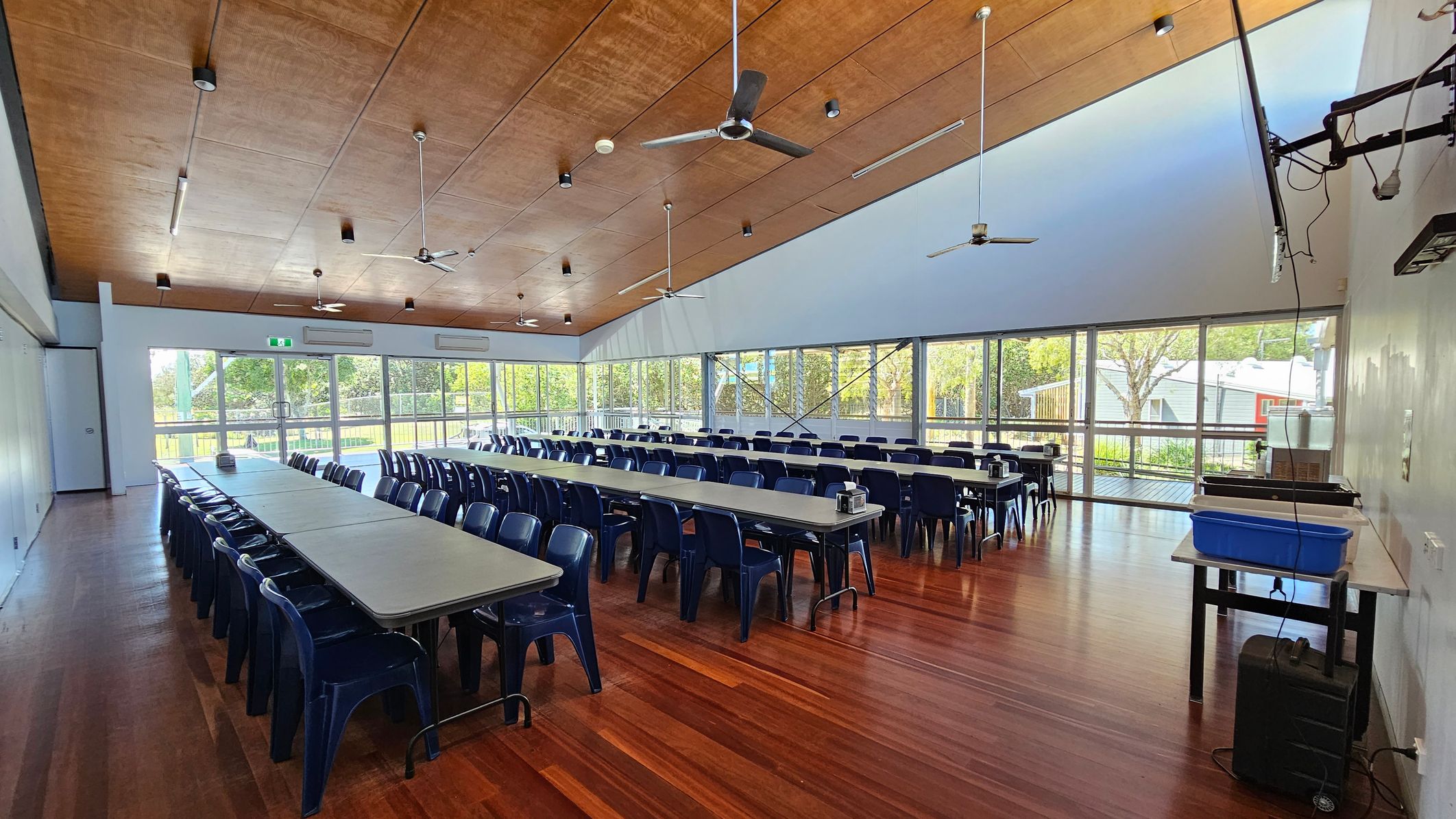 A spacious room with wooden flooring and a wooden ceiling featuring ceiling fans and lights. Long tables with blue chairs are arranged in rows, filling the room. Large windows on the right side let in natural light and offer a view of trees and a building outside. On the right, a table holds a blue container and some equipment. The room has a clean, organised appearance, suitable for meetings or dining. A spacious room with wooden flooring and a wooden ceiling featuring ceiling fans and lights. Long tables with blue chairs are arranged in rows, filling the room. Large windows on the right side let in natural light and offer a view of trees and a building outside. On the right, a table holds a blue container and some equipment. The room has a clean, organised appearance, suitable for meetings or dining.