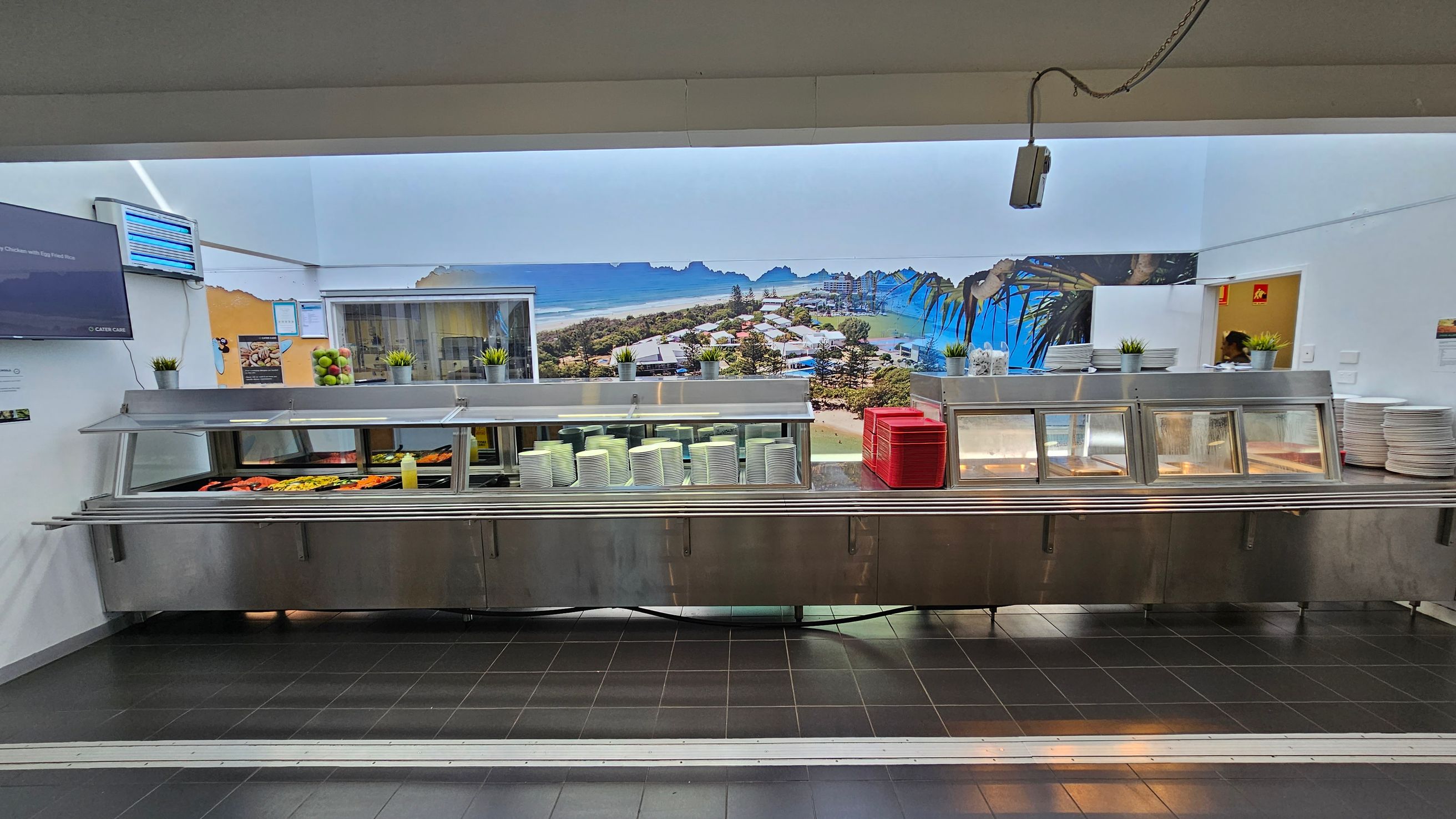 A cafeteria counter with a stainless steel serving area. On the left, there are trays of food, including vegetables and a bottle of sauce. In the centre, stacks of white plates and red trays are visible. On the right, more plates are stacked. Above the counter, small potted plants are placed. The background features a large mural of a coastal landscape with buildings and trees. The floor is tiled in dark grey, and a TV screen is mounted on the left wall. A cafeteria counter with a stainless steel serving area. On the left, there are trays of food, including vegetables and a bottle of sauce. In the centre, stacks of white plates and red trays are visible. On the right, more plates are stacked. Above the counter, small potted plants are placed. The background features a large mural of a coastal landscape with buildings and trees. The floor is tiled in dark grey, and a TV screen is mounted on the left wall.