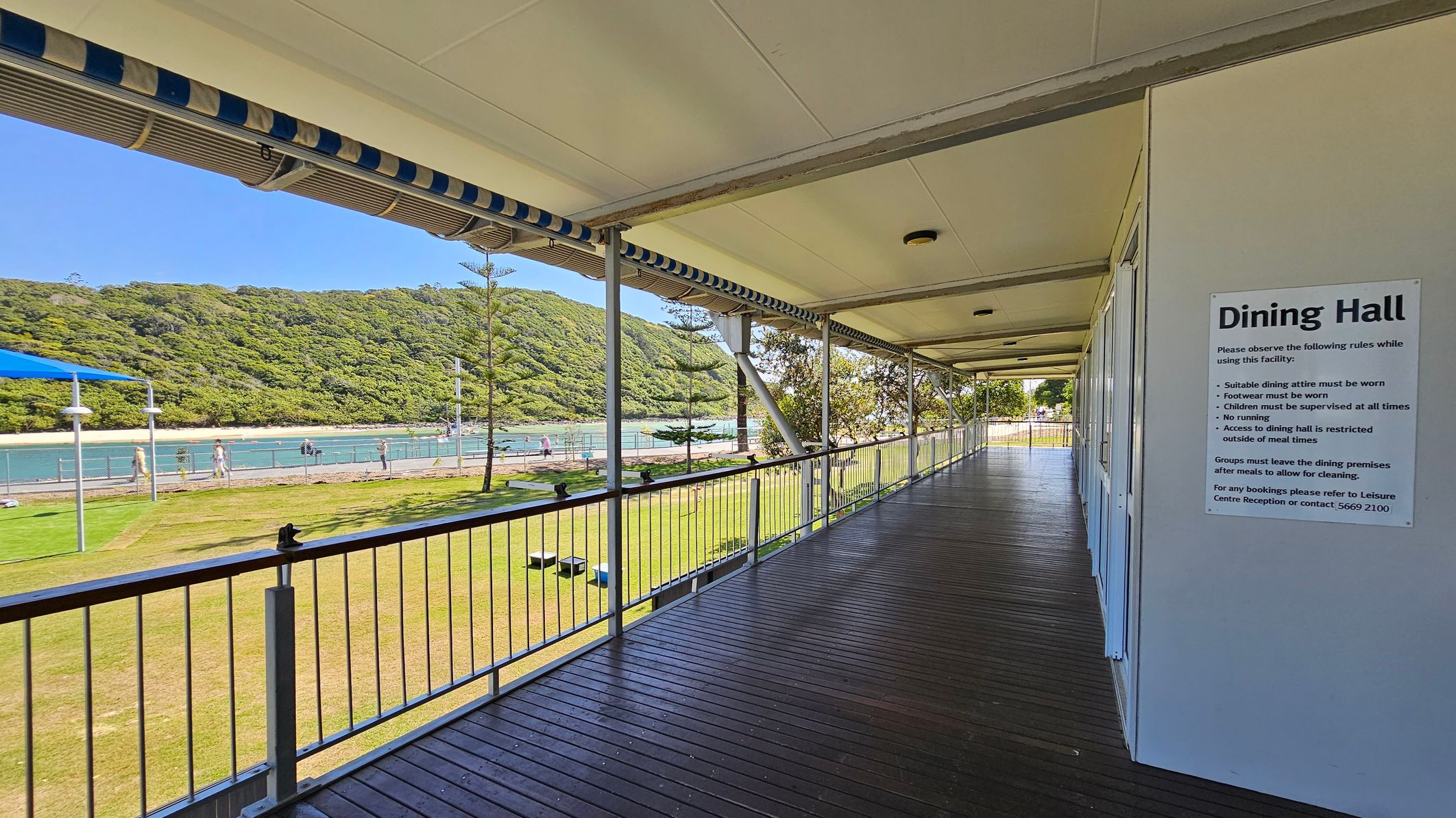 A covered walkway with a wooden floor and a white ceiling runs along the right side, featuring a sign for the Dining Hall with rules. On the left, a railing overlooks a grassy area with a few trees and a blue sunshade. Beyond the grass, there's a body of water with a fence and people walking nearby. In the background, a lush, green hill rises, providing a natural backdrop. A covered walkway with a wooden floor and a white ceiling runs along the right side, featuring a sign for the Dining Hall with rules. On the left, a railing overlooks a grassy area with a few trees and a blue sunshade. Beyond the grass, there's a body of water with a fence and people walking nearby. In the background, a lush, green hill rises, providing a natural backdrop.