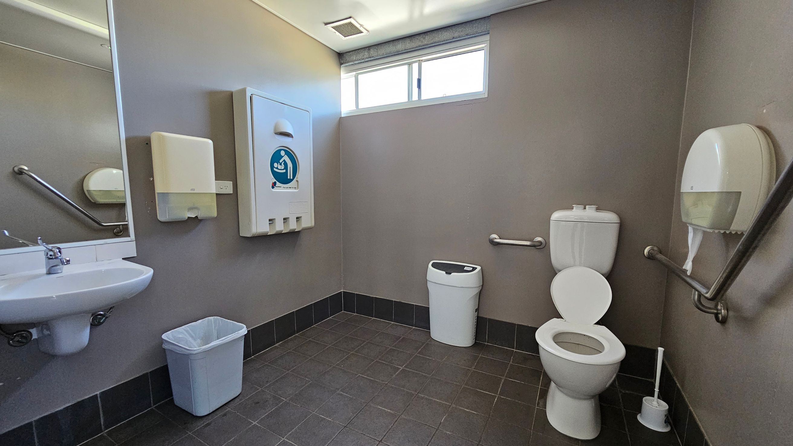 A public toilet with grey walls and a tiled floor. On the left, there's a white sink with a mirror above it and a paper towel dispenser. Below the sink is a blue waste bin. In the centre, a baby changing station is mounted on the wall. On the right, a white toilet with grab bars on either side and a toilet brush nearby. Above the toilet is a small window, and a toilet paper dispenser is mounted on the wall. A public toilet with grey walls and a tiled floor. On the left, there's a white sink with a mirror above it and a paper towel dispenser. Below the sink is a blue waste bin. In the centre, a baby changing station is mounted on the wall. On the right, a white toilet with grab bars on either side and a toilet brush nearby. Above the toilet is a small window, and a toilet paper dispenser is mounted on the wall.
