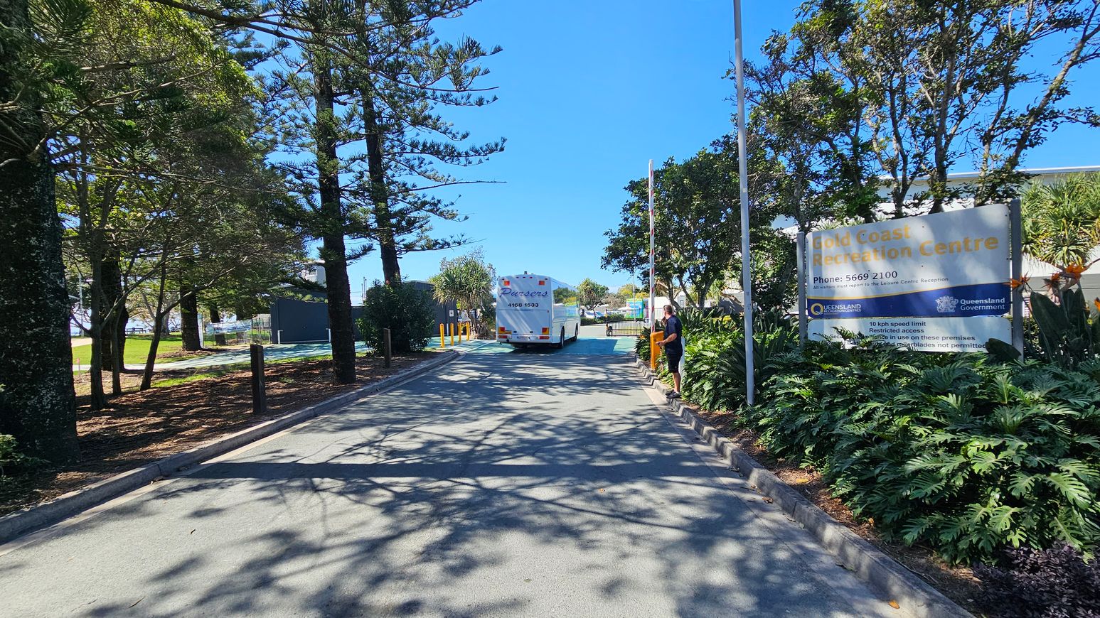 A road lined with tall trees on the left, casting shadows on the pavement. On the right, a large sign reads "Gold Coast Recreation Centre" with contact information and rules. A person stands near the sign. In the centre, a white bus is driving away, with greenery and more trees in the background. The scene is well-lit, suggesting a sunny day. A road lined with tall trees on the left, casting shadows on the pavement. On the right, a large sign reads "Gold Coast Recreation Centre" with contact information and rules. A person stands near the sign. In the centre, a white bus is driving away, with greenery and more trees in the background. The scene is well-lit, suggesting a sunny day.