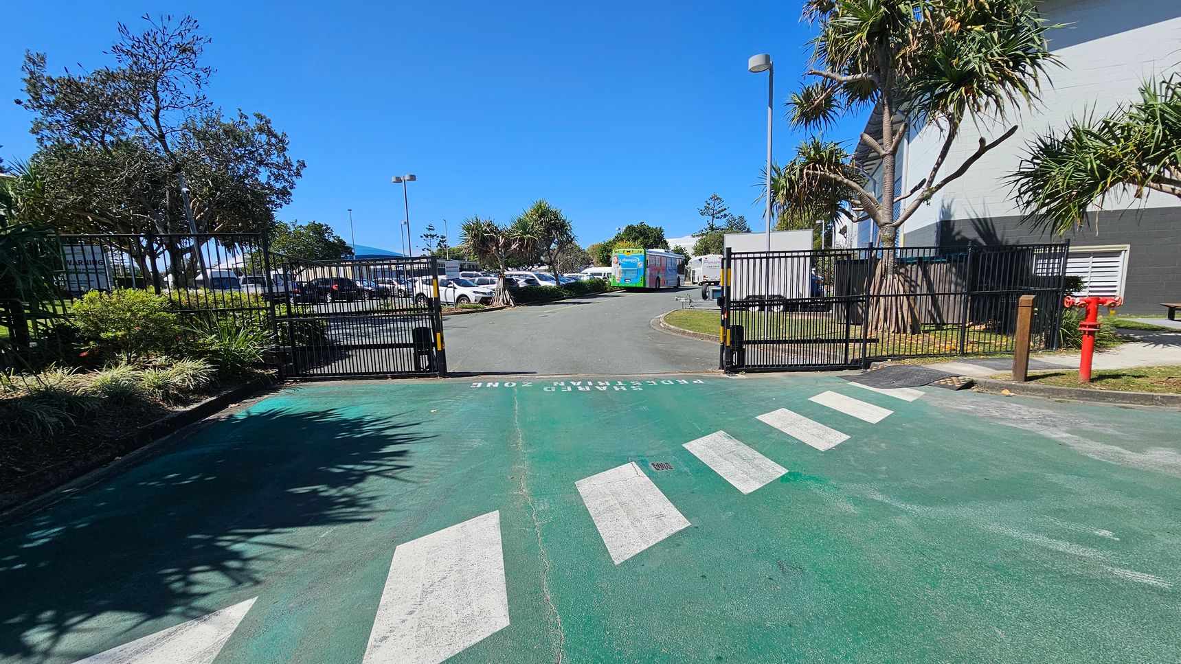 A gated entrance with a green pedestrian zone and white zebra crossing in the foreground. The gate is open, leading to a car park with several vehicles and a colourful bus in the background. On the left, there are trees and shrubs, while on the right, a building with a grey and white exterior and a red fire hydrant is visible. The sky is clear and blue. A gated entrance with a green pedestrian zone and white zebra crossing in the foreground. The gate is open, leading to a car park with several vehicles and a colourful bus in the background. On the left, there are trees and shrubs, while on the right, a building with a grey and white exterior and a red fire hydrant is visible. The sky is clear and blue.