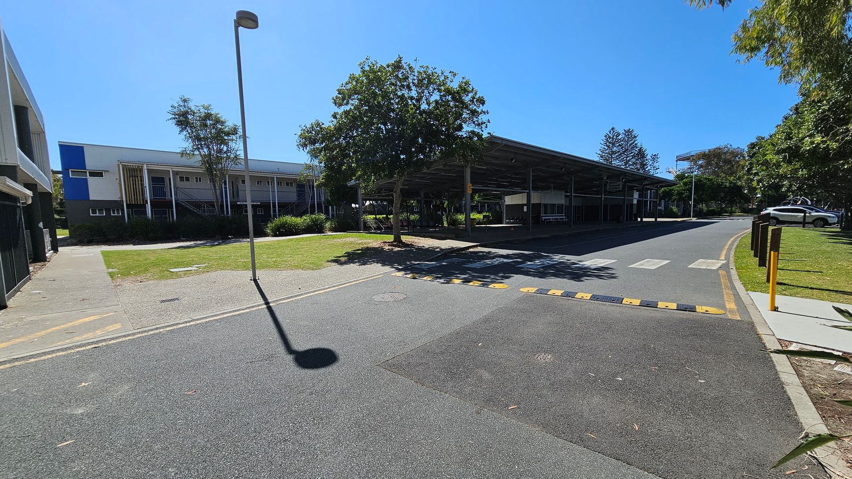 A paved road with a speed bump and a pedestrian crossing runs through the centre. On the left, there's a building with a blue and white facade, and a grassy area with a tree. On the right, a covered structure with benches and another tree is visible. A car is parked further down the road. The area is surrounded by greenery, and a tall streetlamp casts a shadow on the road. A paved road with a speed bump and a pedestrian crossing runs through the centre. On the left, there's a building with a blue and white facade, and a grassy area with a tree. On the right, a covered structure with benches and another tree is visible. A car is parked further down the road. The area is surrounded by greenery, and a tall streetlamp casts a shadow on the road.