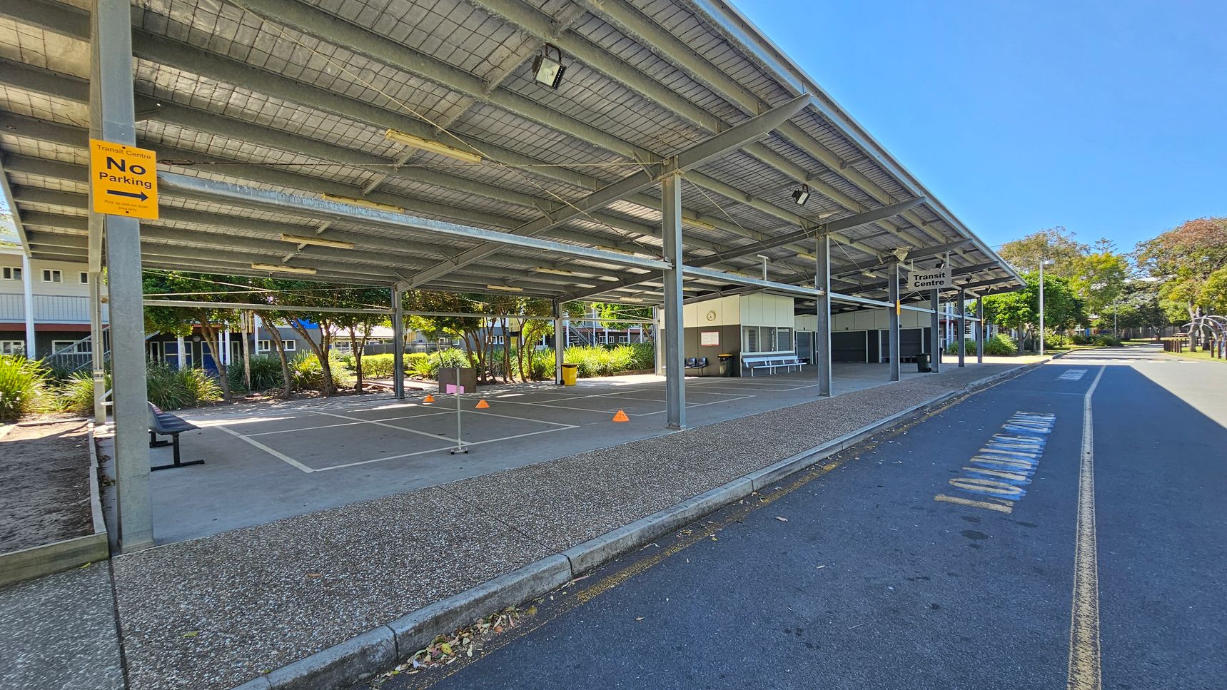 A covered area with a metal roof and support beams, featuring a "No Parking" sign on the left. The ground is paved with a speckled texture. There are marked parking spaces with orange cones and a bench. In the background, there are buildings with windows and greenery, including trees and shrubs. On the right, a road with faded markings runs alongside the structure, and a "Transit Centre" sign is visible. The area is well-lit with natural light. A covered area with a metal roof and support beams, featuring a "No Parking" sign on the left. The ground is paved with a speckled texture. There are marked parking spaces with orange cones and a bench. In the background, there are buildings with windows and greenery, including trees and shrubs. On the right, a road with faded markings runs alongside the structure, and a "Transit Centre" sign is visible. The area is well-lit with natural light.