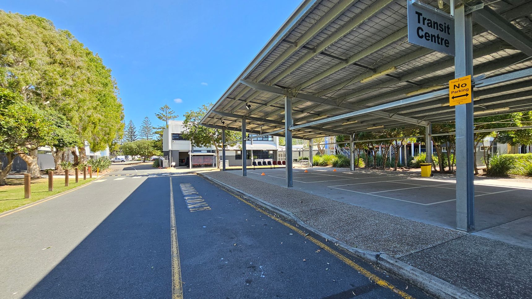 A transit centre with a large metal roof structure on the right, providing shade over a paved area. A sign reads "Transit Centre" with a "No Parking" notice below it. On the left, a road with a marked lane runs alongside a row of trees and grass. In the background, there are buildings with trees and shrubs around them. The ground is a mix of asphalt and concrete, and the area is well-lit. A transit centre with a large metal roof structure on the right, providing shade over a paved area. A sign reads "Transit Centre" with a "No Parking" notice below it. On the left, a road with a marked lane runs alongside a row of trees and grass. In the background, there are buildings with trees and shrubs around them. The ground is a mix of asphalt and concrete, and the area is well-lit.