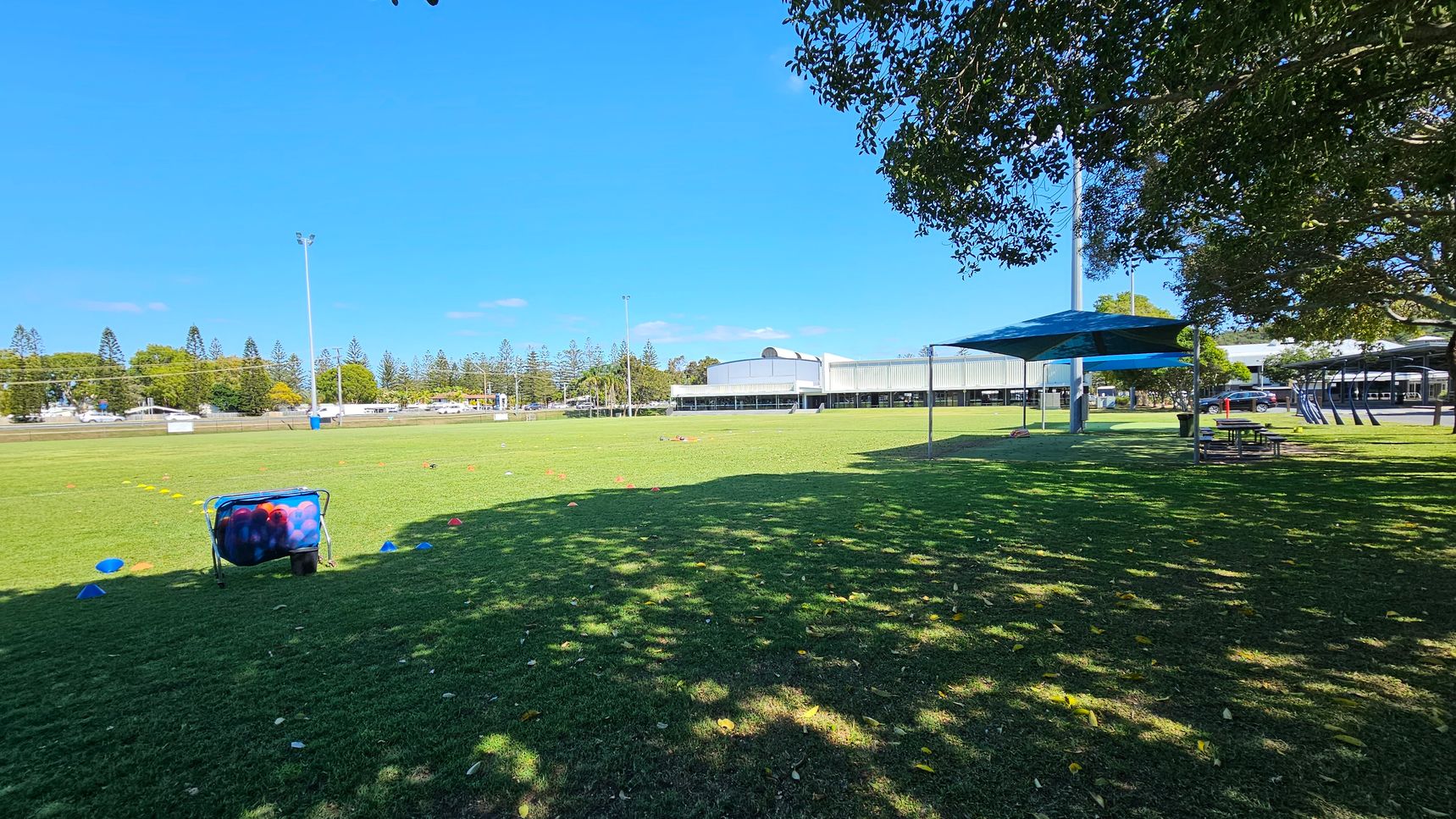 A grassy field with scattered sports cones and a basket of balls on the left. In the background, there are tall trees and a large building. On the right, a shaded area with picnic tables under a canopy. The foreground shows shadows from nearby trees. A grassy field with scattered sports cones and a basket of balls on the left. In the background, there are tall trees and a large building. On the right, a shaded area with picnic tables under a canopy. The foreground shows shadows from nearby trees.