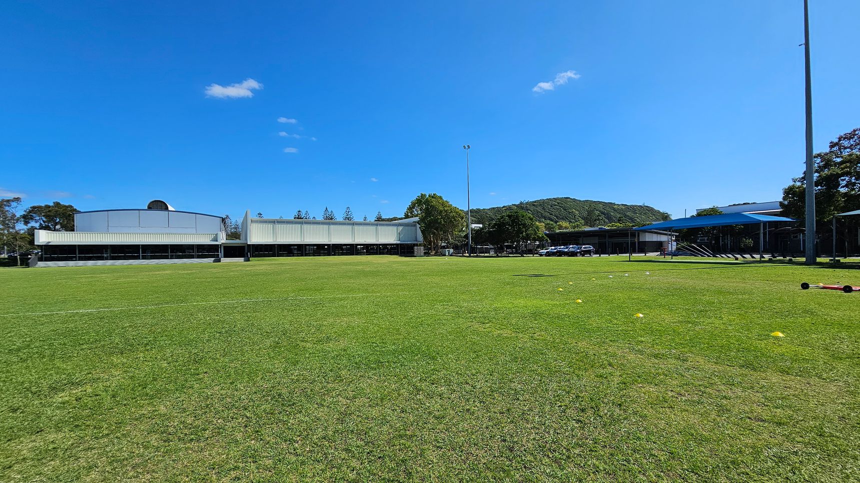 A large grassy field with a few scattered yellow cones in the foreground. In the background, there are several modern buildings with white facades. To the right, a covered area with a blue roof is visible, along with a row of parked cars. A tall light pole stands near the centre, and trees are visible behind the buildings, with hills in the distance. The sky is clear with a few small clouds. A large grassy field with a few scattered yellow cones in the foreground. In the background, there are several modern buildings with white facades. To the right, a covered area with a blue roof is visible, along with a row of parked cars. A tall light pole stands near the centre, and trees are visible behind the buildings, with hills in the distance. The sky is clear with a few small clouds.