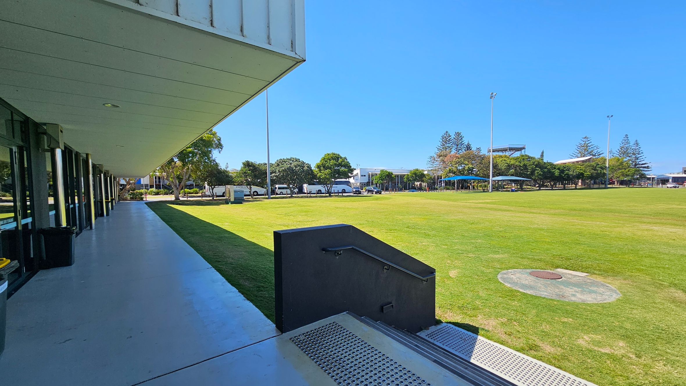 A wide, open grassy field is on the right, bordered by trees and several tall light poles. In the background, there are buildings and a row of parked vehicles. On the left, a modern building with large glass windows and a covered walkway extends along the edge of the field. The foreground features a set of steps with a metal railing leading down to the grass. A wide, open grassy field is on the right, bordered by trees and several tall light poles. In the background, there are buildings and a row of parked vehicles. On the left, a modern building with large glass windows and a covered walkway extends along the edge of the field. The foreground features a set of steps with a metal railing leading down to the grass.