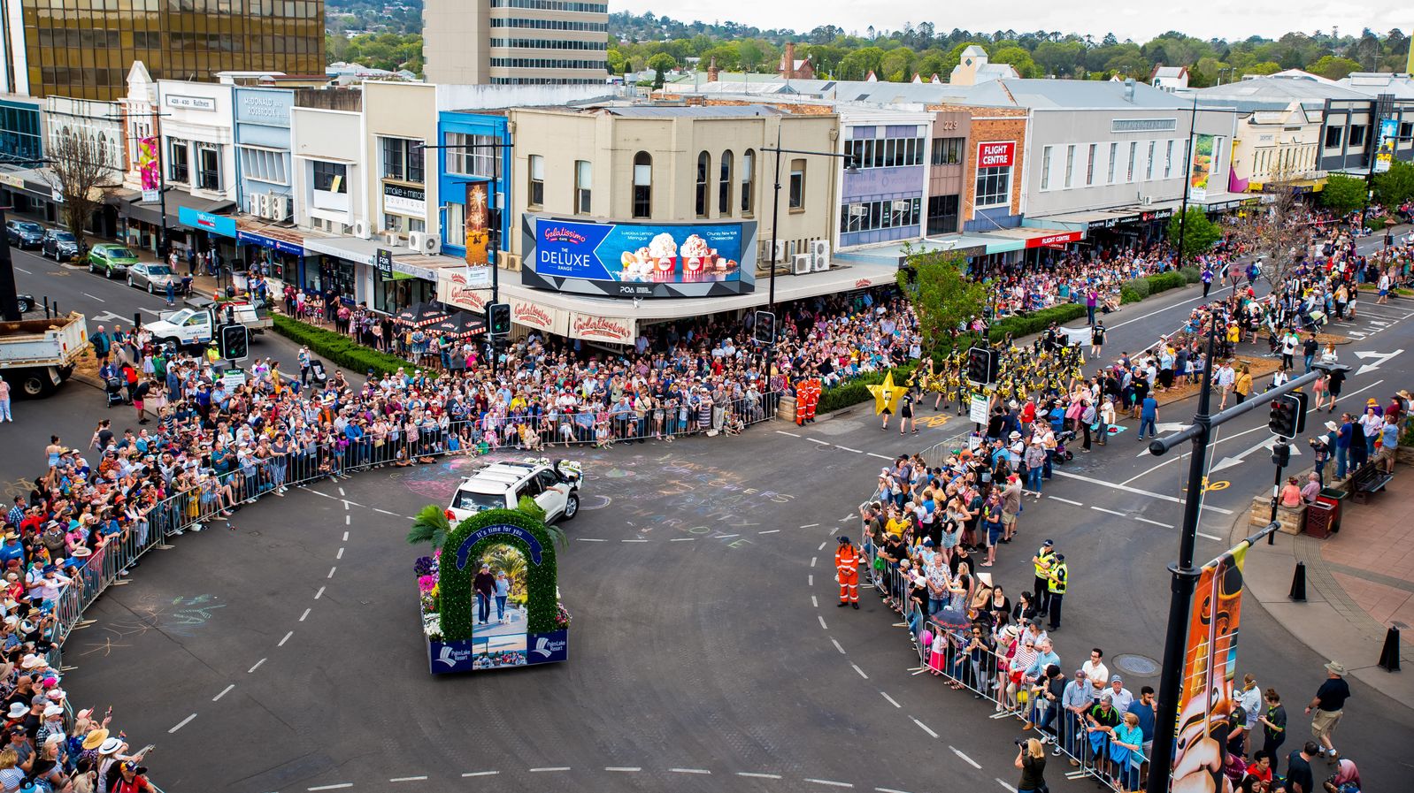 A bustling street parade with a large crowd gathered on both sides of the road. In the centre, a white vehicle with a decorative arch on its back is moving along the street. The crowd is contained by metal barriers, and many people are taking photos. Buildings with various shopfronts and advertisements line the background. A group of performers in costumes is visible on the right, and a few police officers are present for crowd control.