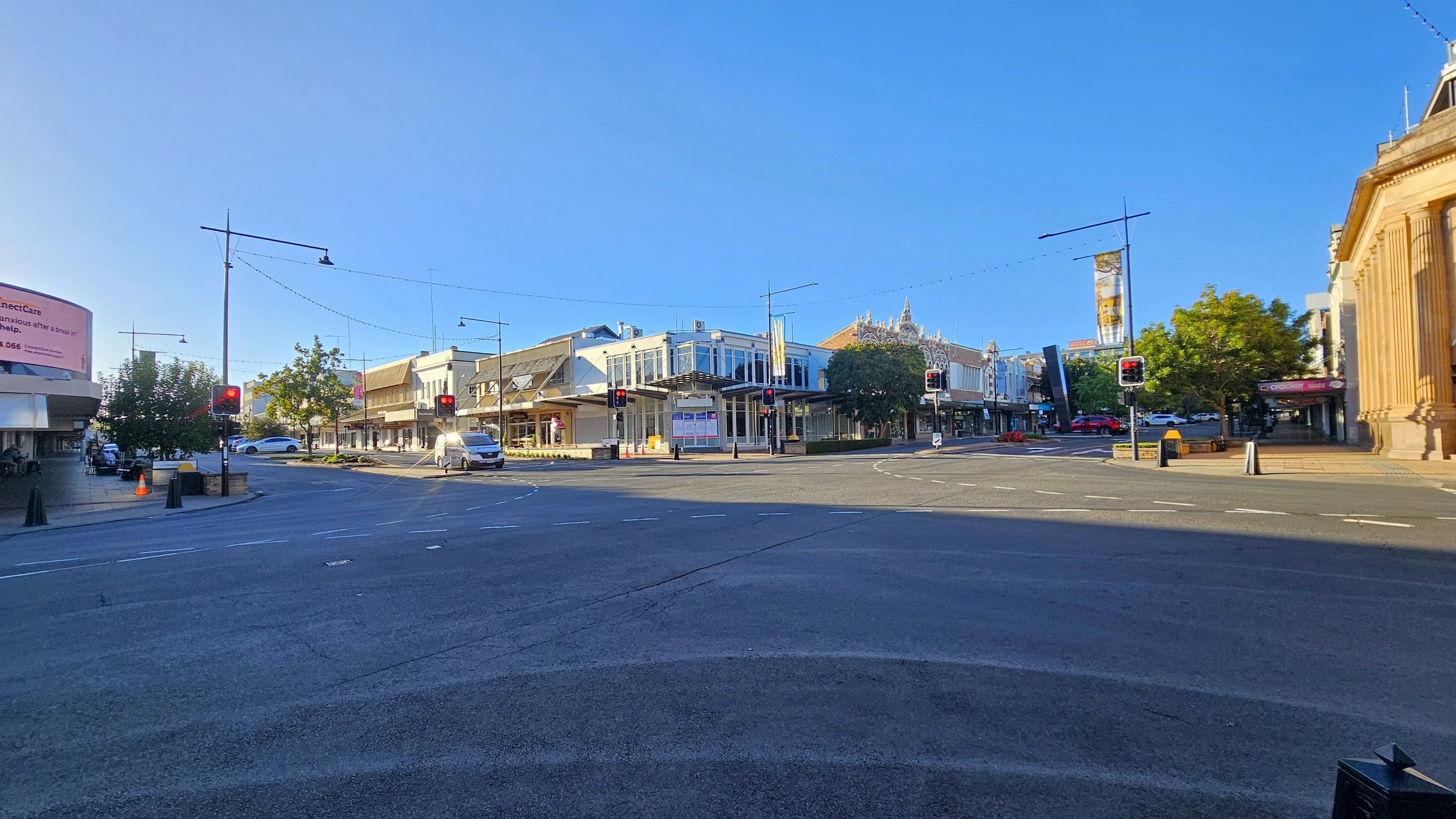 A street intersection with traffic lights on either side. On the left, there's a large billboard and a few trees. In the centre, a row of buildings with glass windows and decorative facades. A white van and a few cars are visible on the road. On the right, a building with tall columns. The road surface is dark, and the sky is clear.