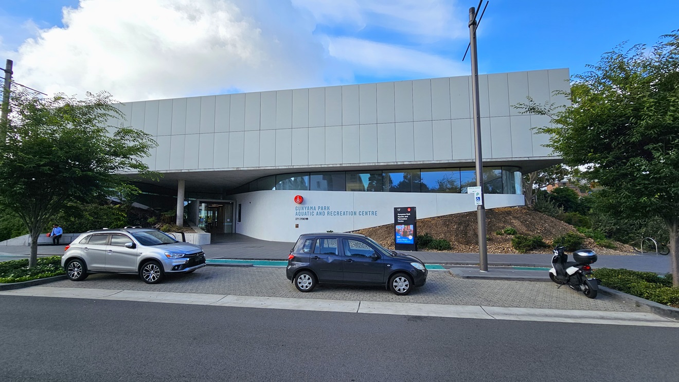 In the centre stands a commercial building crafted from concrete, glass, and tiles. The facility's name is displayed on the building's facade. Adjacent to the building, a car park accommodates two cars and a motorcycle. Landscape concrete plant boxes flank both sides of the car park. On the right, an electric post and a large banner are situated near the sloping hillside of the road. A bike rack is also present.