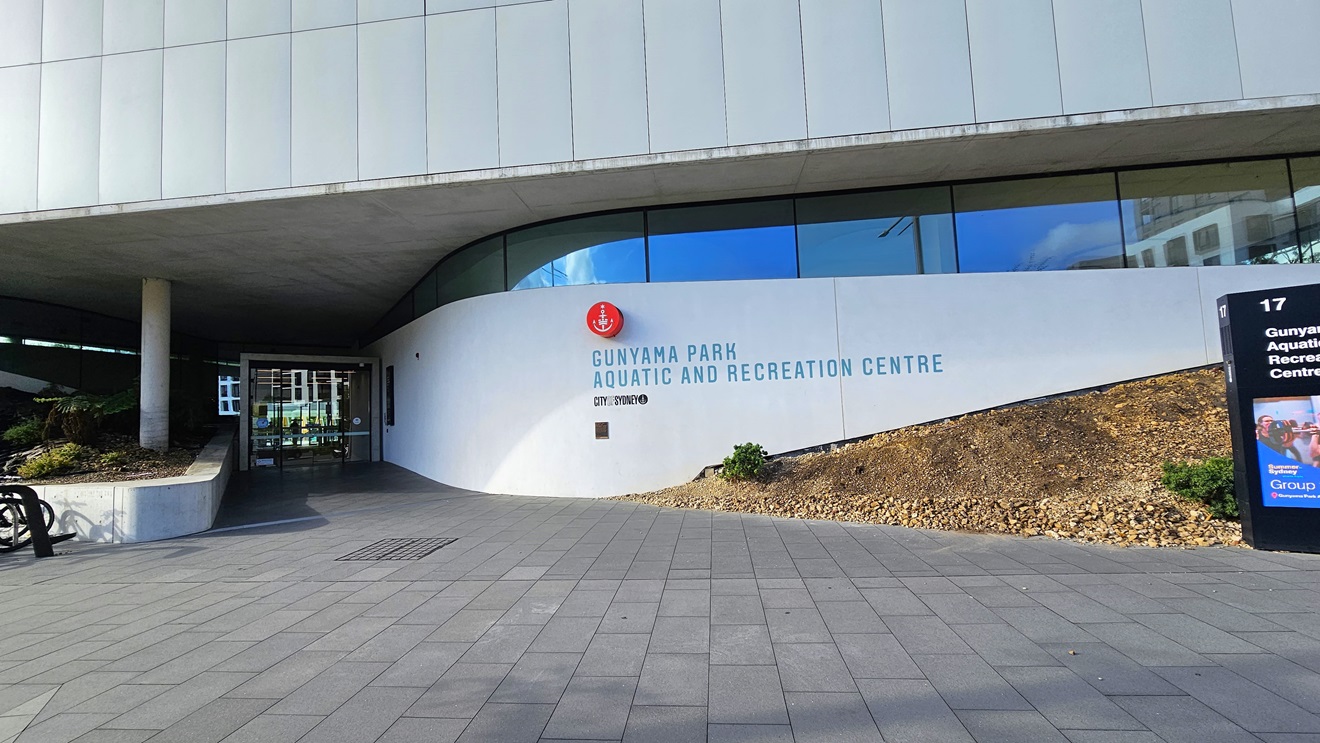 At the centre is a commercial building constructed with concrete, glass, and tiles. The facility's name is shown on the right facade, alongside another large signage. The entrance, on the left, features a floor-to-ceiling glass door. A concrete plant box with a landscape ornamental plant is positioned on the left. The floor is covered with concrete tiles.