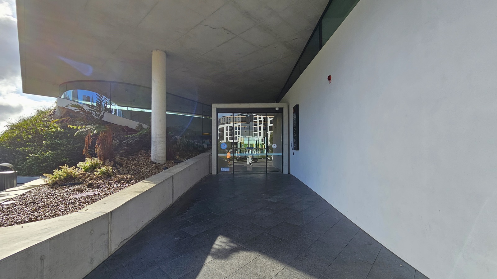 To the left, a concrete plant box holds landscape ornamental plants and pebbles. A large concrete post supports the ceiling. The right wall is constructed from white concrete. In the middle, the entrance showcases a floor-to-ceiling automatic glass door. The floor is surfaced with concrete tiles, and the ceiling is white.