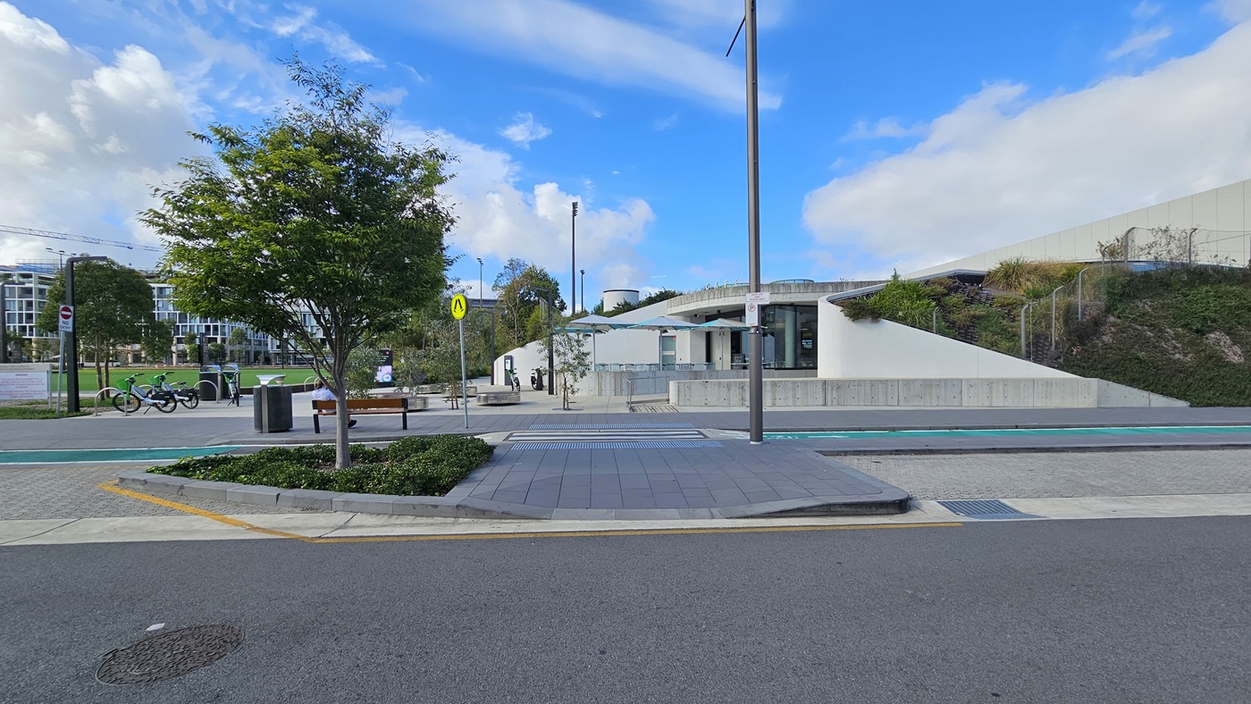 An exterior view of the commercial building reveals a structure crafted from concrete and glass, offering shaded seating in front. The car park includes a step-free ramp and a pedestrian lane leading to the building. A concrete plant box showcases ornamental plants and trees, accompanied by a bench. Bike racks and bicycles are situated to the left. On the right, a hillside features wild plants and grass. A tall commercial building and green grass are visible on the far left.