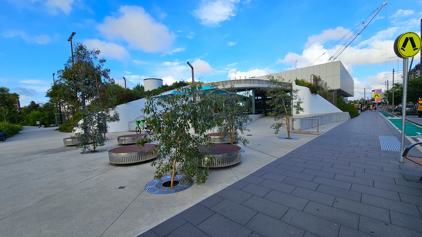 At the centre stands a white commercial building, constructed from concrete, glass, and tiles. The building's concourse includes round concrete seating and small trees. To the right, the road is equipped with road signs, a step-free ramp, and truncated domes. Cars are visible in the vicinity.