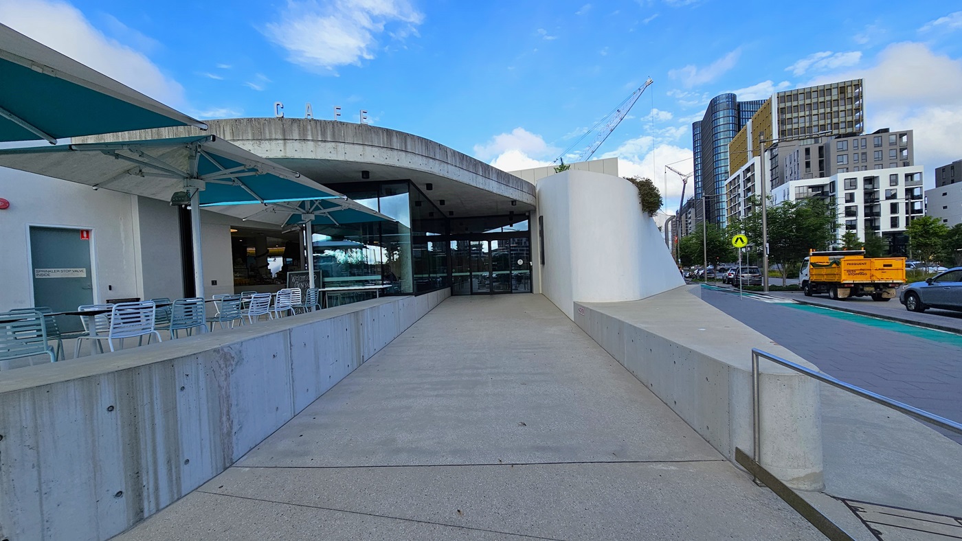 In the centre is a white commercial building, made of concrete, glass, and tiles, with bold "Cafe" letters affixed to the roof. On the left, umbrella shades cover arranged tables and chairs. A ramp concrete pathway leads to the building's entrance, featuring floor-to-ceiling glass. On the right, a road accommodates various vehicles, with tall commercial buildings and trees visible.