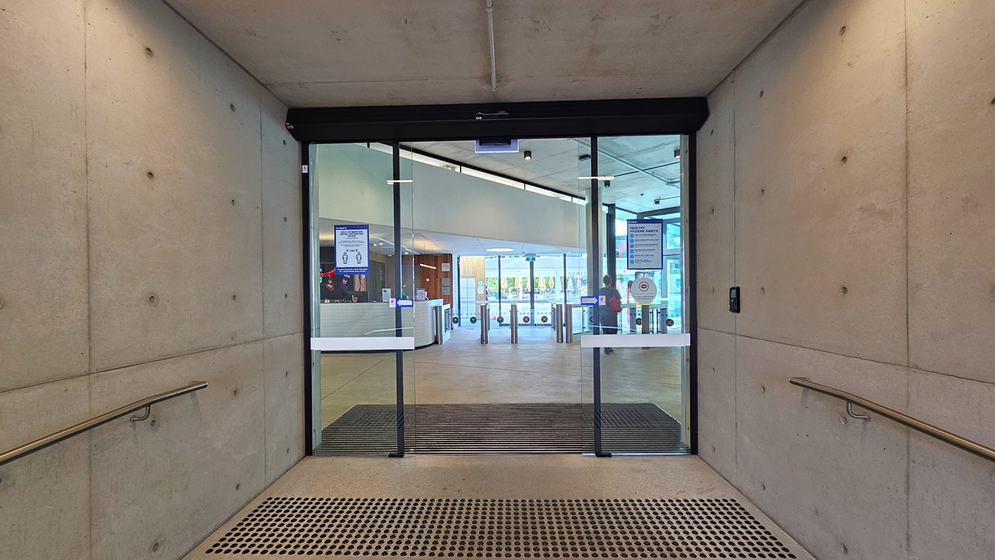 The entrance features a floor-to-ceiling automatic glass door with truncated domes and an anti-slip mat. Posters are affixed to the glass wall. Railings are present on both sides of the white wall. Inside, the reception desk and a controlled gate are visible on the left.