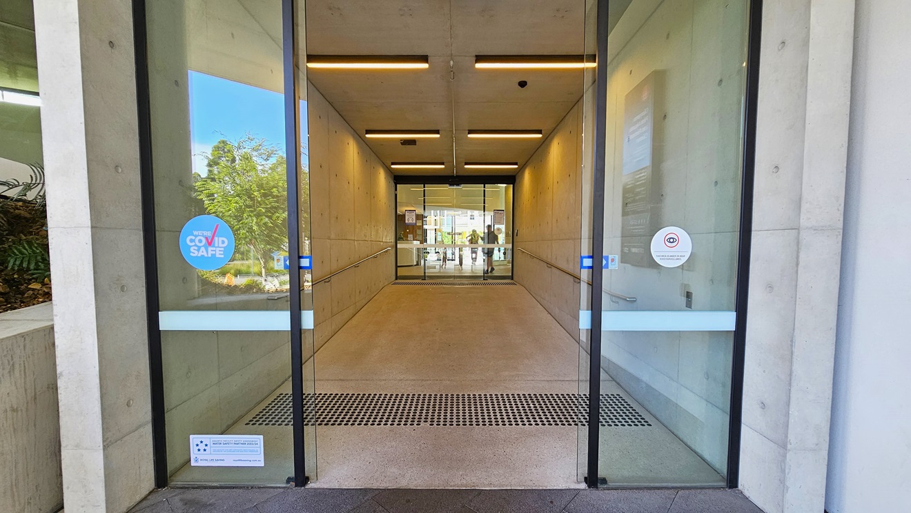 The entrance is equipped with double automatic floor-to-ceiling glass doors featuring truncated domes for safety. Safety signs and sticker reminders are affixed to the glass wall. The lobby displays white walls with a poster on the right, and the ceiling is white with fluorescent lights. Behind the second glass door, the reception area is visible.