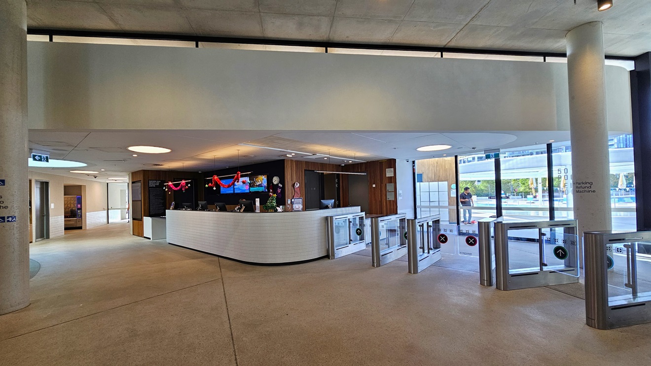The reception desk is a white counter with a brick design. On the countertop and ceiling are computers and Christmas decorations. The reception wall has a dark wooden design. On the left, a lobby leads to other facilities. The floor is concrete, and the ceiling is white with LED lights. To the right, turnstiles marked for separate entry and exit are present, with the Aquatic facility also visible.