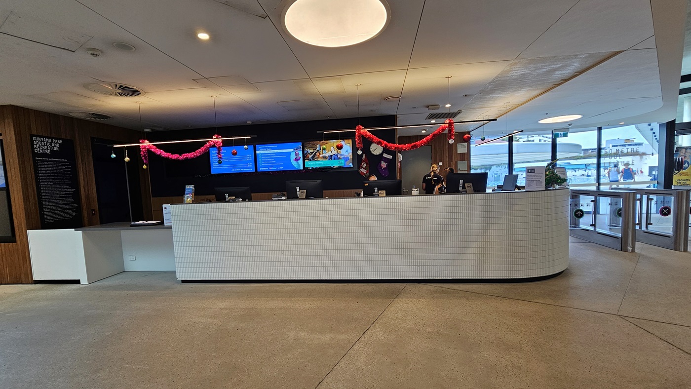 The reception desk is a white counter with a brick design, featuring a lower counter on the left. Computers and Christmas decorations adorn the countertop and ceiling. The reception wall has a dark wooden design, with flat-screen TVs mounted on it. The floor is concrete, and the ceiling is white with LED lights. To the right, turnstiles marked for separate entry and exit are present, with the Aquatic facility also visible.