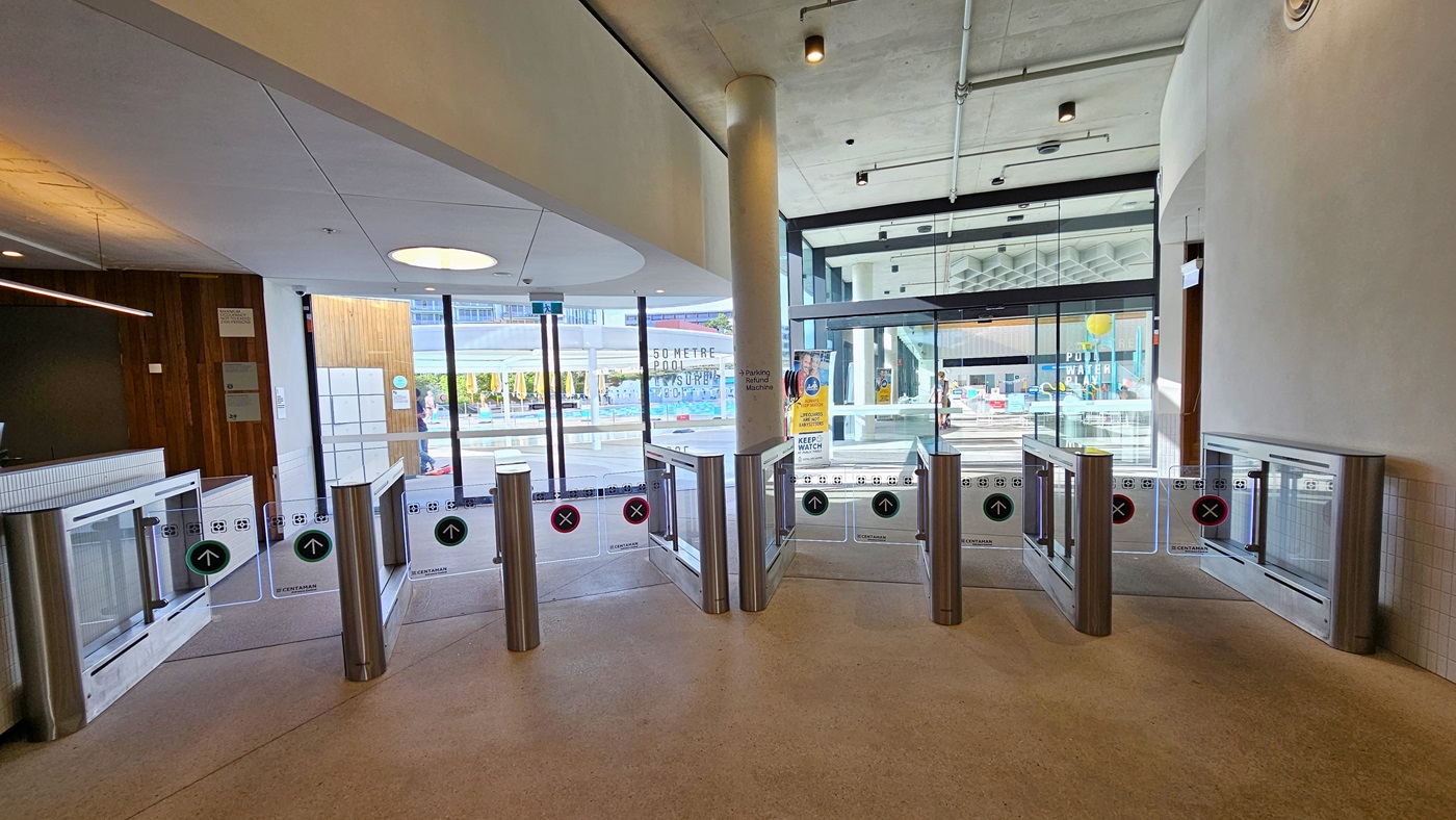 In the centre are glass turnstiles marked for separate entry and exit. Beyond the gates, another glass entrance door leads to the Aquatic Centre. On the left, a brown wall displays posters, while the right wall is white. The ceiling is white with pin lights and LED lights. The floor is constructed of concrete.