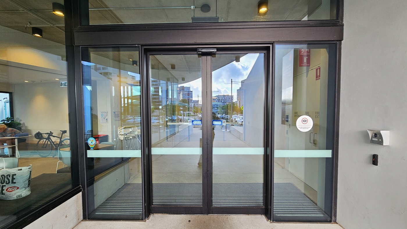 In the middle is a floor-to-ceiling automatic glass sliding door with anti-slip mats. Posted stickers are affixed to the glass wall. An alcohol dispenser is mounted on the right wall. To the left, a glass wall encloses seating inside. The right wall is uniformly white, and the ceiling is white as well.