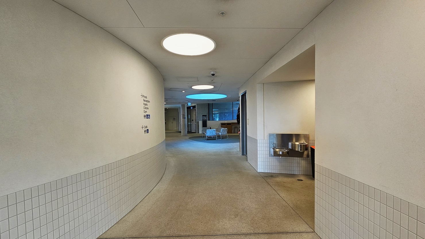 A corridor with white walls with small white tile accents. Directional signs are affixed to the left wall. On the right, a drinking fountain is situated. Further, a seating area is arranged with movable chairs and tables. The ceiling is white with large round LED lights, and the floor is made of concrete.
