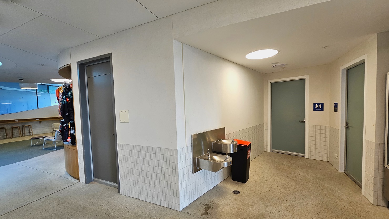 To the right, a drinking fountain is positioned next to a rubbish bin. Two grey doors, each with gender access labels, are in view. On the left, another grey door is present. Tables and chairs, along with retail items, are visible on the left. The ceiling is white with large round LED lights, and the floor is made of concrete. The walls feature white with small white tiles for accent.