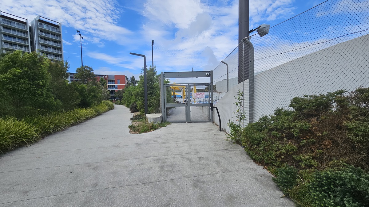 To the left, a tall commercial building, trees, and ornamental plants border the road. In the middle, a concrete path leads to a gate with a glass door. To the right, a white wall with a chain fence is present. The hillside section on the right is covered with wild grass and shrubs.