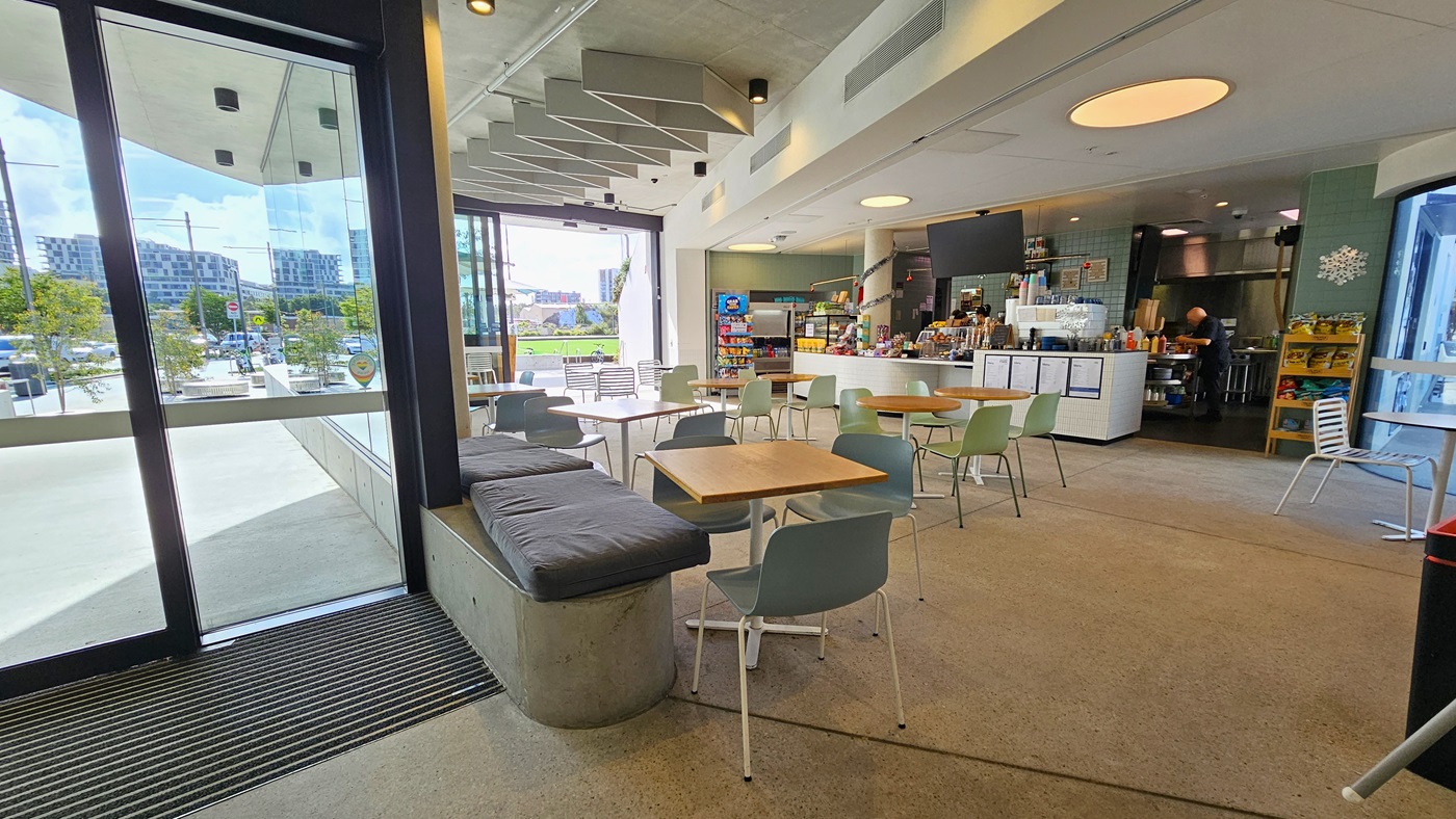 The glass door entrance features a non-slip mat. Inside the cafe, movable tables and chairs are arranged. To the right, the reception desk has a white brick counter equipped with posters. Food items are displayed on the countertop. A flat-screen TV is mounted to the ceiling. Display racks showcasing chips and light snacks are positioned on both sides of the counter. The ceiling features a diamond-shaped wooden design and has large circular LED lights. A Glass wall is visible on the left.