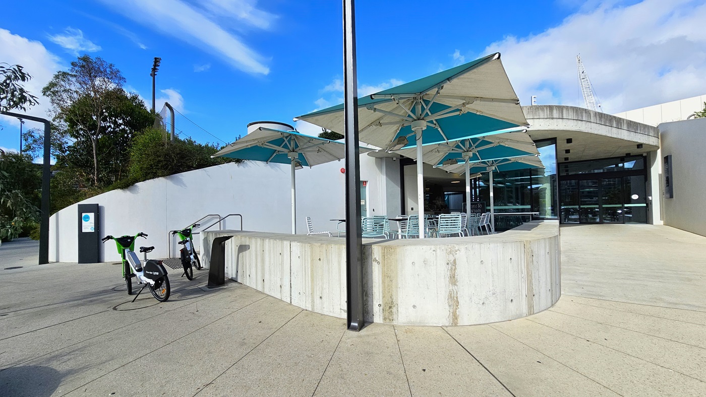 The concrete ramp curves and elevates, leading to the glass door entrance. To the left, large umbrella shades are positioned in front of the cafe. A stepped entry, equipped with a handrail, is located on the left, with two bicycles parked nearby.