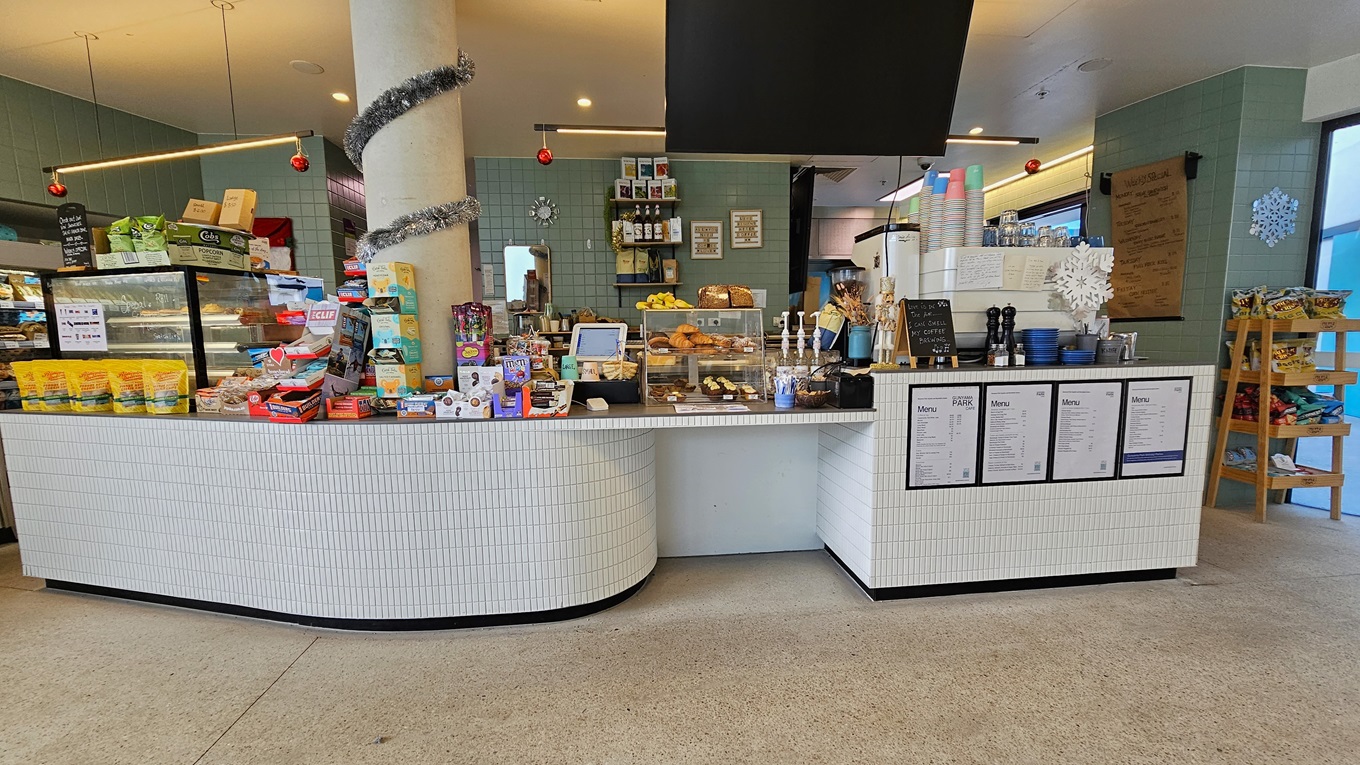 The cafe's reception counter is adorned with small white tiles. Food racks, snacks, and treats are displayed on the countertop. The left section of the counter holds four posters with the menu, drinks dispenser, and plastic cups. A display rack showcases light snacks on the right. A flat-screen TV is mounted from the ceiling. Christmas decorations adorn the space. The floor is covered with speckled grey tiles, and the ceiling and walls are white and light green, respectively.